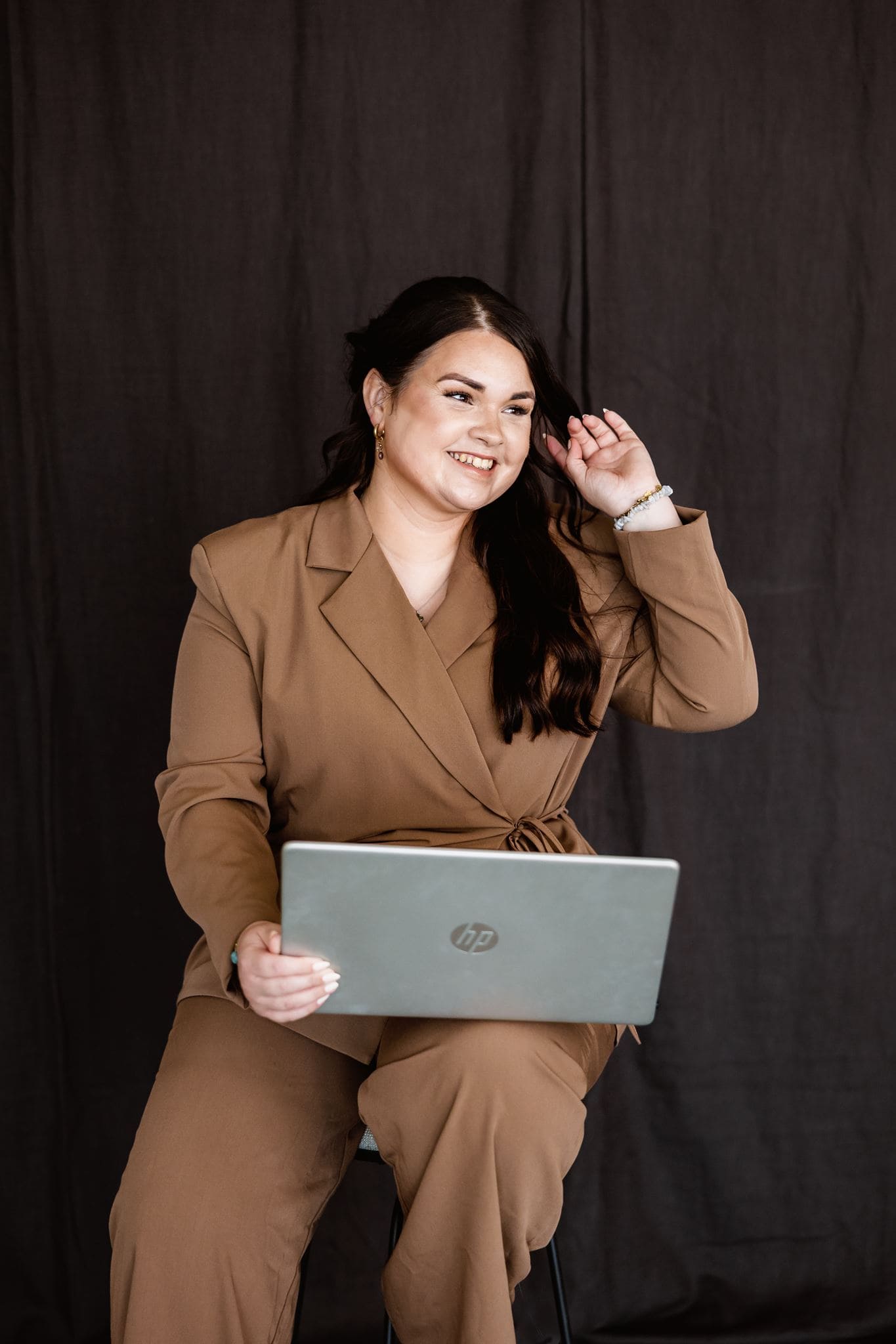 Een wedding planner in een bruin pak zit met een laptop op haar schoot; ze glimlacht en strijkt haar haar naar achteren tijdens een fotoshoot in de studio.