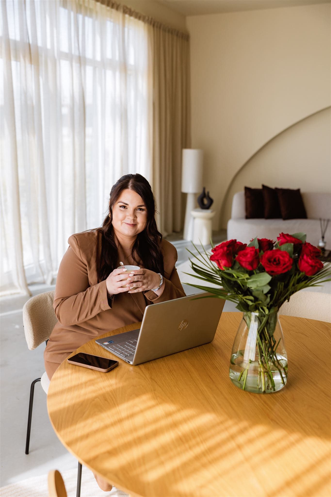 Weddingplanner zit tijdens haar brandingshoot aan een tafel met laptop en boeket met rode rozen.
