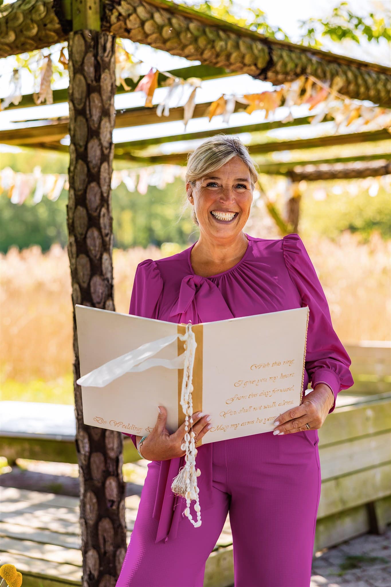 Glimlachende BABS in een felroze outfit houdt een open boek vast buiten onder een houten luifel versierd met linten.