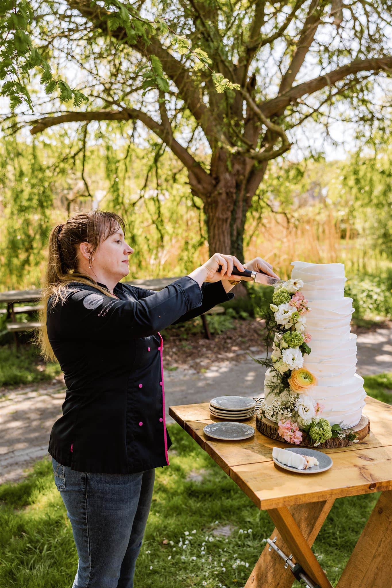 Een vrouw snijdt een hoge, versierde witte taart buiten bij een houten tafel met borden in een tuinsetting tijdens een branding shoot.