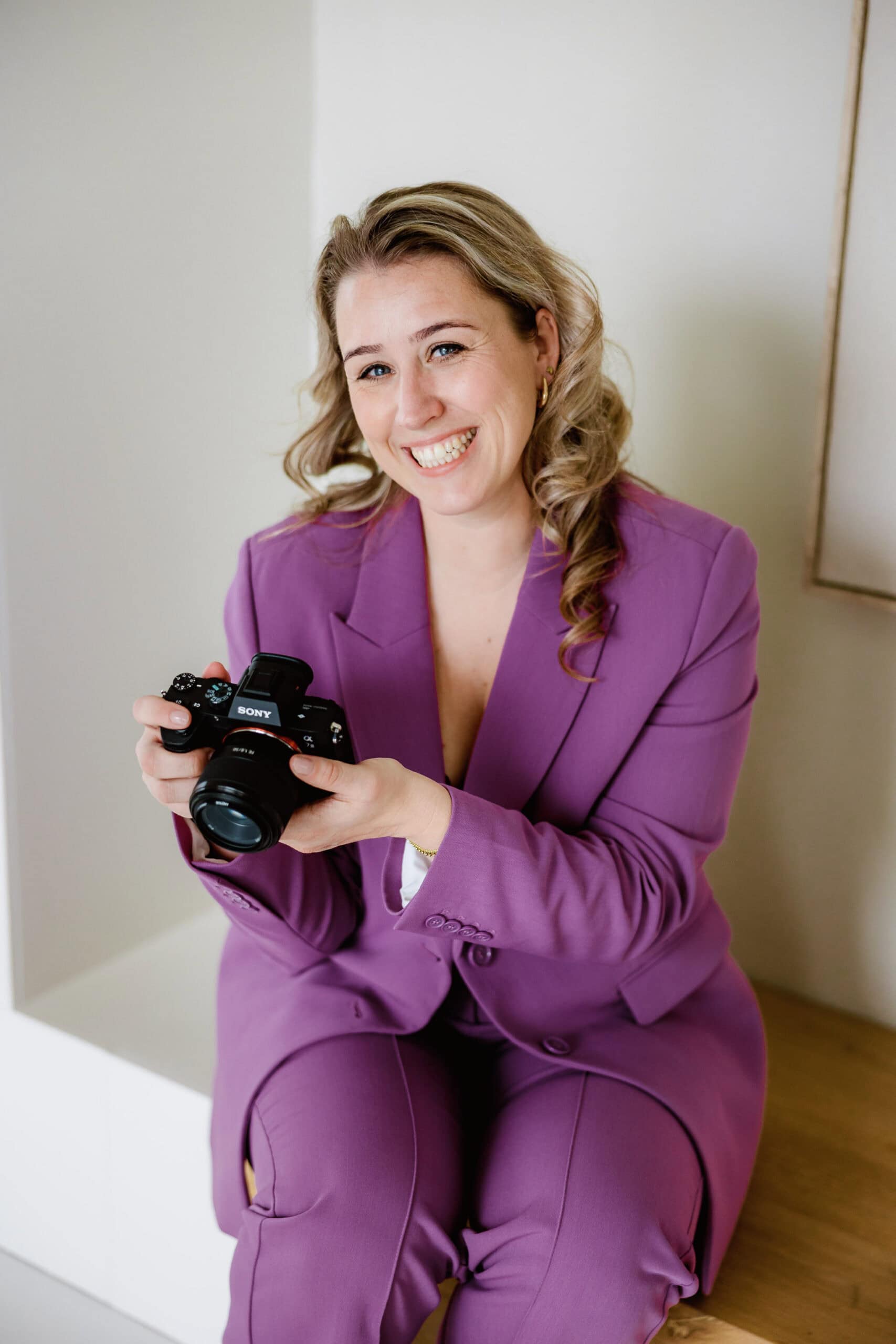 Glimlachende fotografe in een paars pak met camera, zittend binnen tegen een lichte achtergrond tijdens een branding shoot.