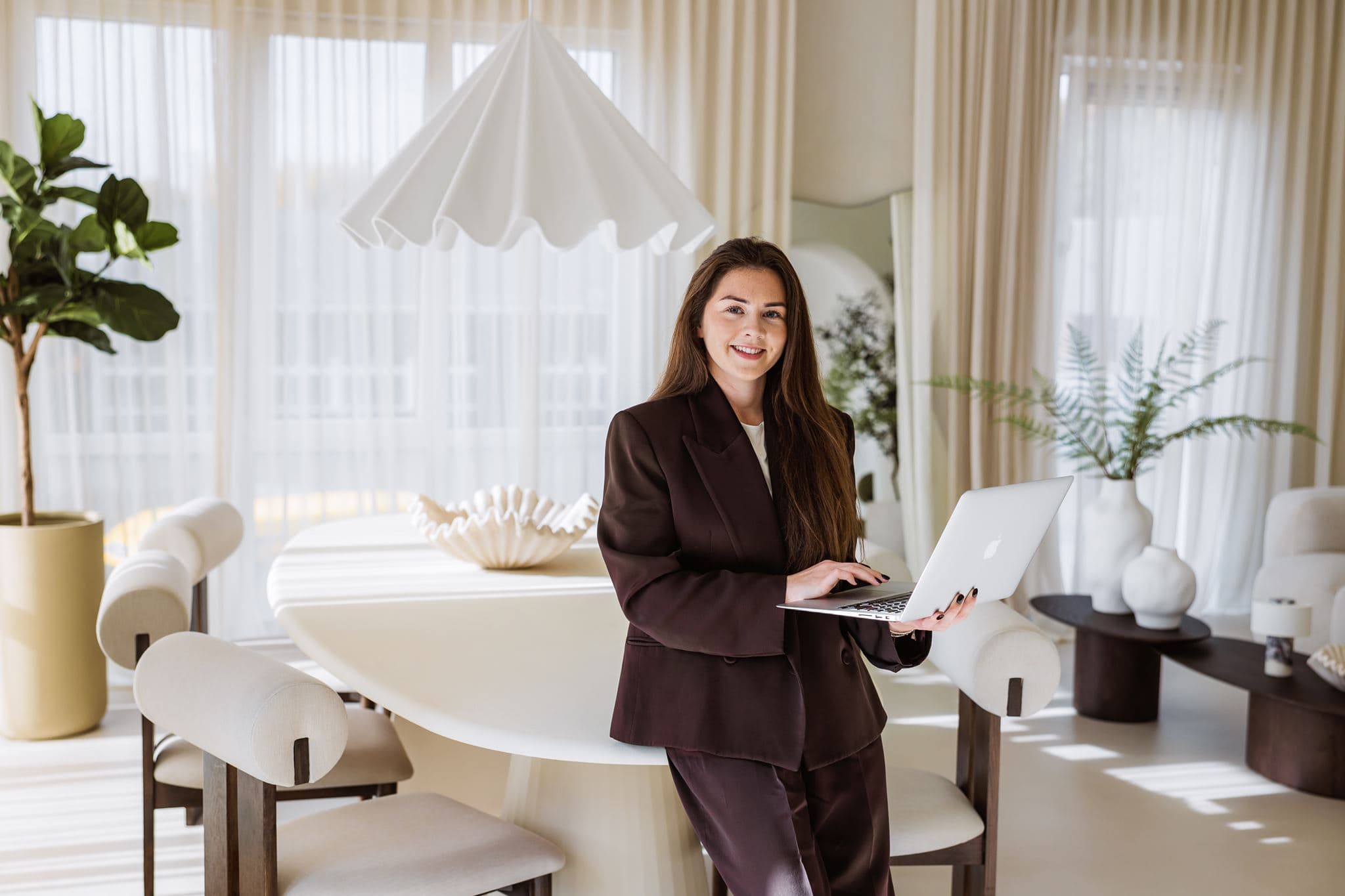 Een brandingfoto van een vrouwelijke weddingplanner in een bruin pak met haar laptop in de hand in een daglichtstudio.