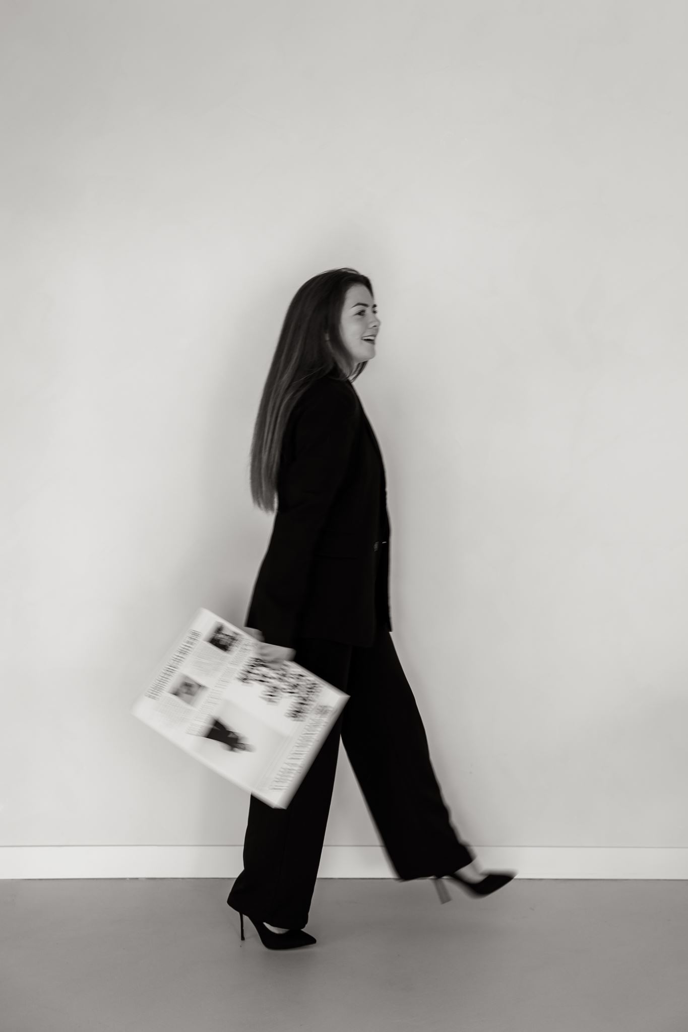 Vrouw in een pak en op hakken, lopend met een krant in haar hand, tegen een effen lichte muur; zwart-witfoto door fotograaf Drenthe, Groningen, Friesland.