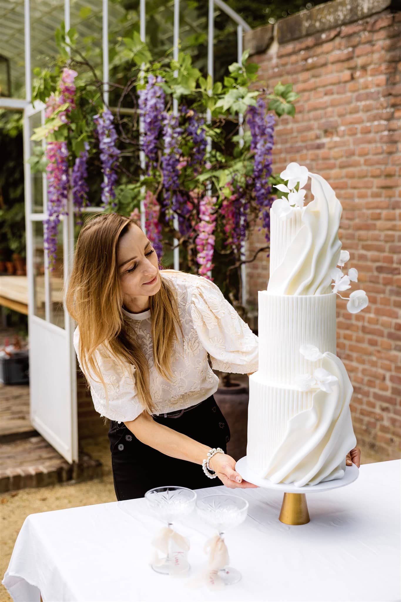 Een vrouw schikt een hoge, elegante witte taart met bloemdecoraties op een buitentafel tijdens een branding shoot.