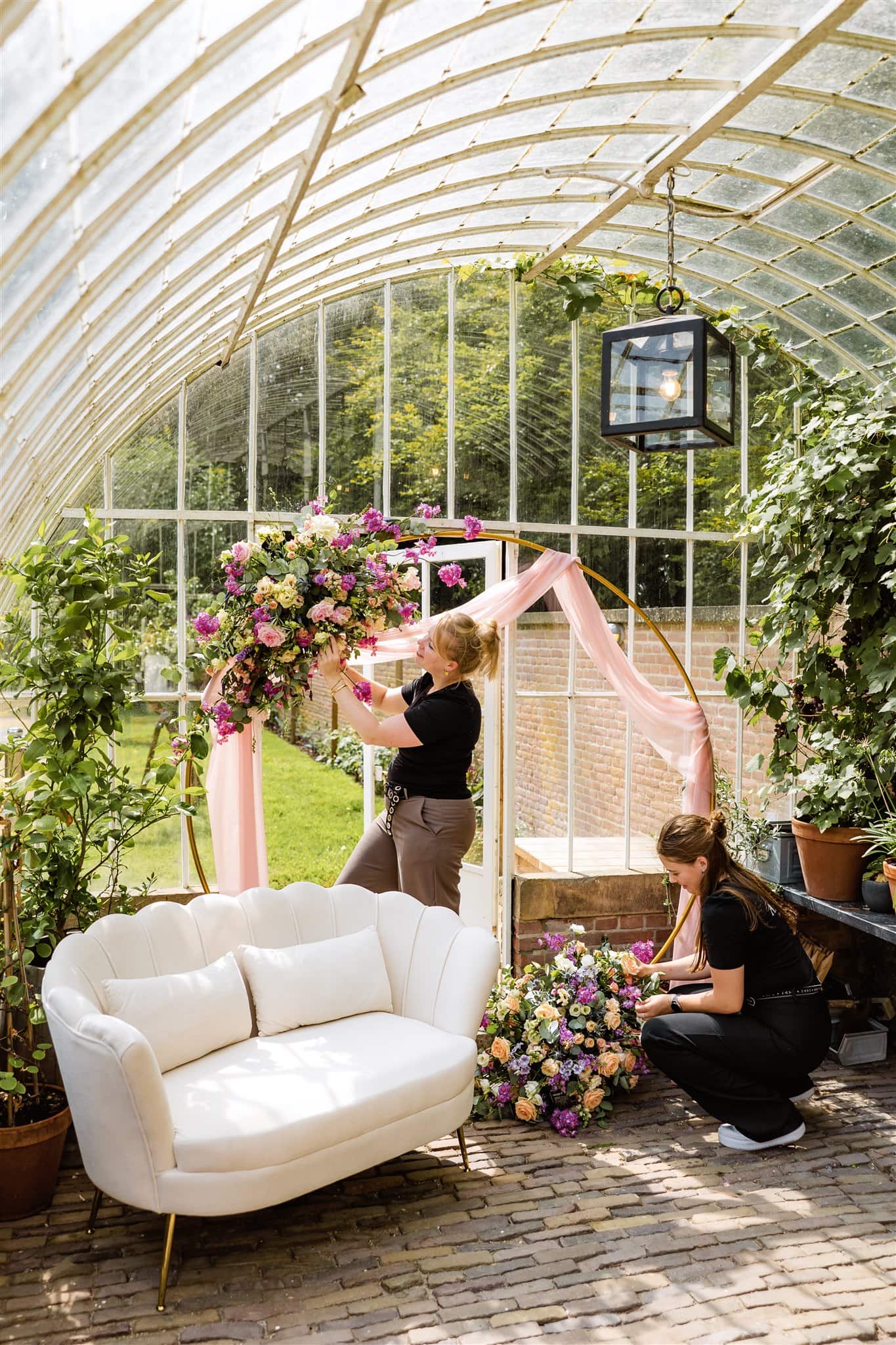 Twee bruidsstylisten versieren een bloemenboog met roze en paarse bloemen in een kas naast een witte bank.