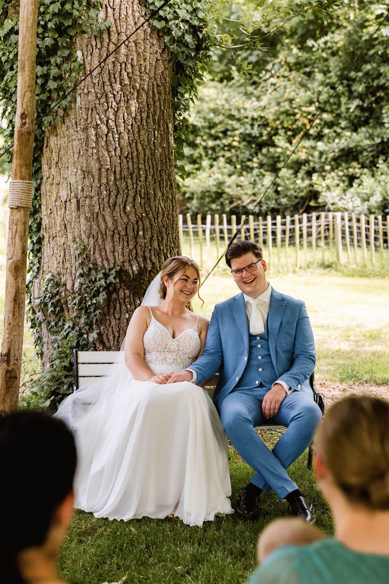 Een bruid en bruidegom zitten samen glimlachend onder een boom tijdens hun trouwceremonie bij Hotel Wesseling in Dwingeloo, vastgelegd door een trouwfotograaf Drenthe.