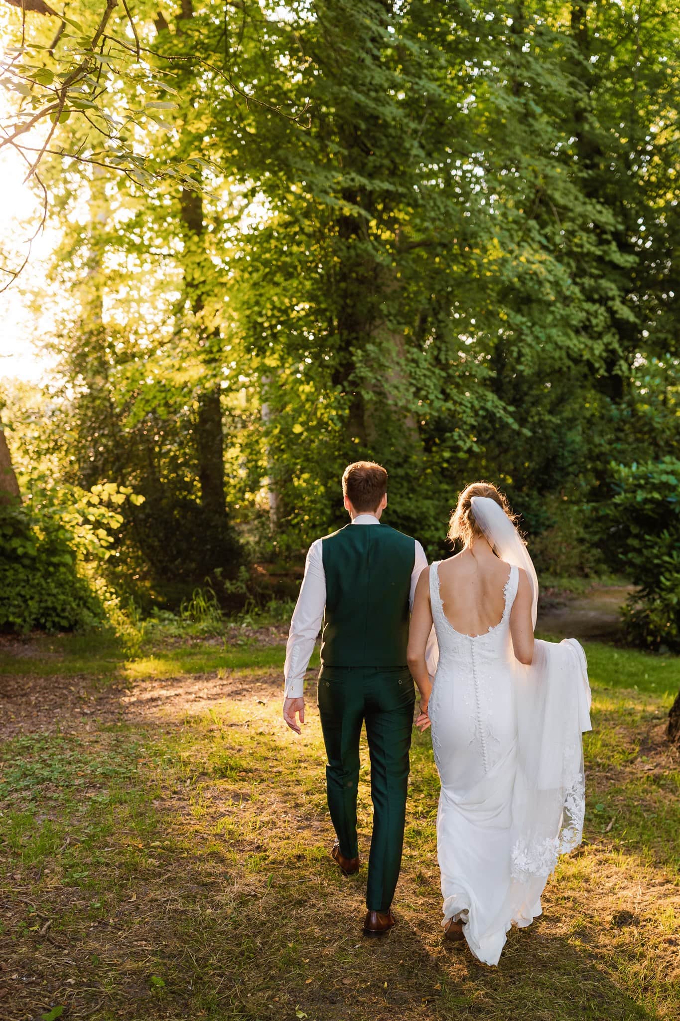 Een bruid en bruidegom lopen hand in hand door een groen bos bij Landgoed Lemferdinge, met golden hour zonsondergang licht.