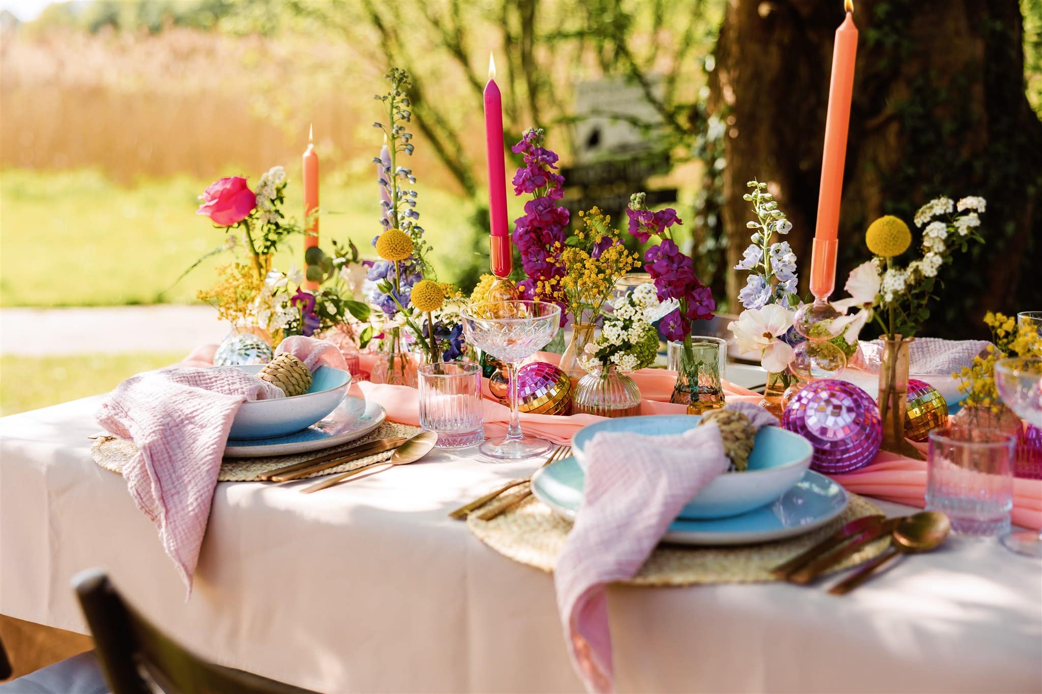 Kleurrijk gedekte tafel op een bruiloft met bloemen, kaarsen, discoballen en pastelkleurig servies op een zonnige dag.