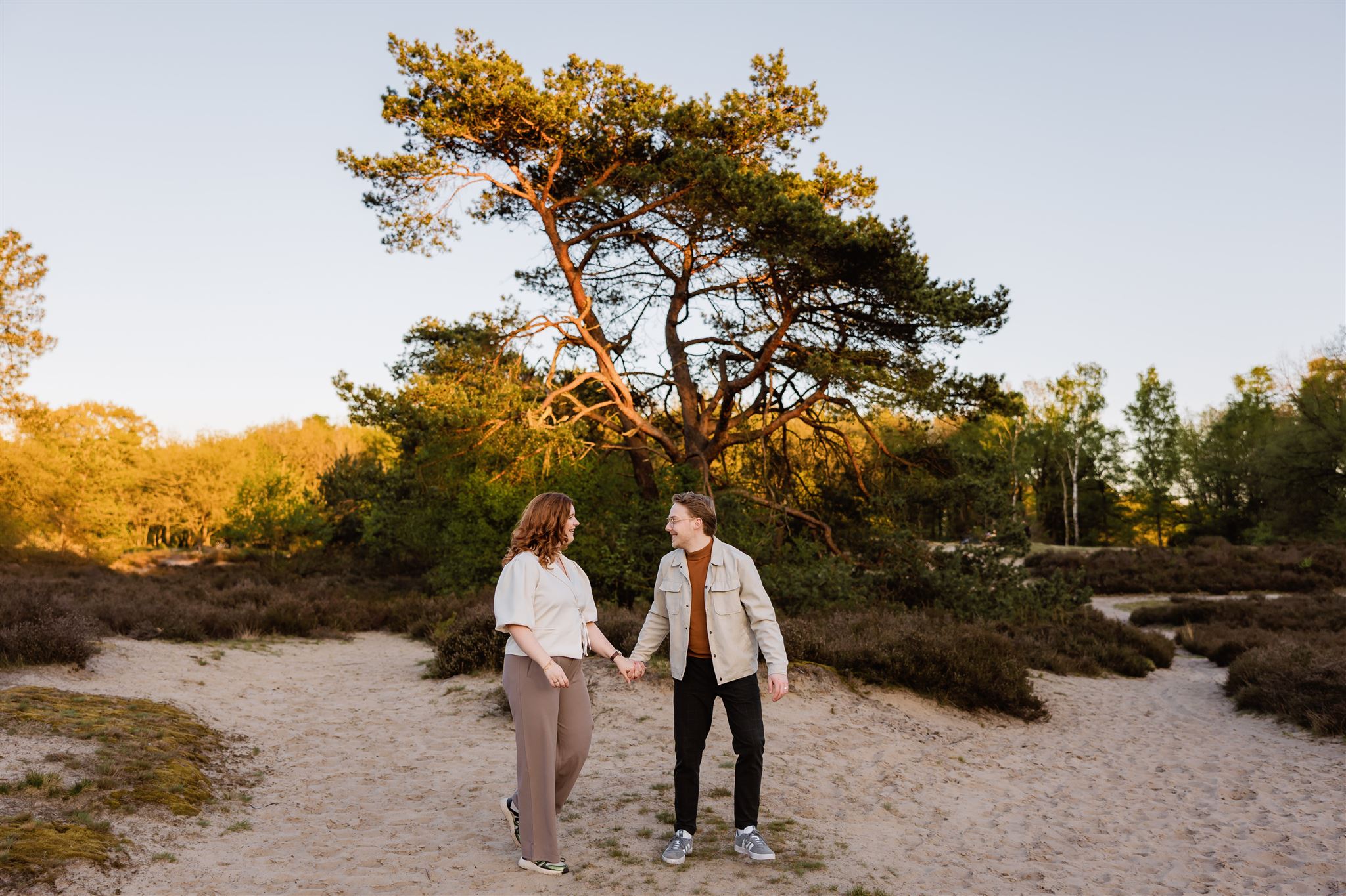 Een paar dat elkaars hand vasthoudt en naar elkaar lacht terwijl ze op zandgrond staan met bomen op de achtergrond, perfect voor iedereen die op zoek is naar kledingtips fotoshoot inspiratie.
