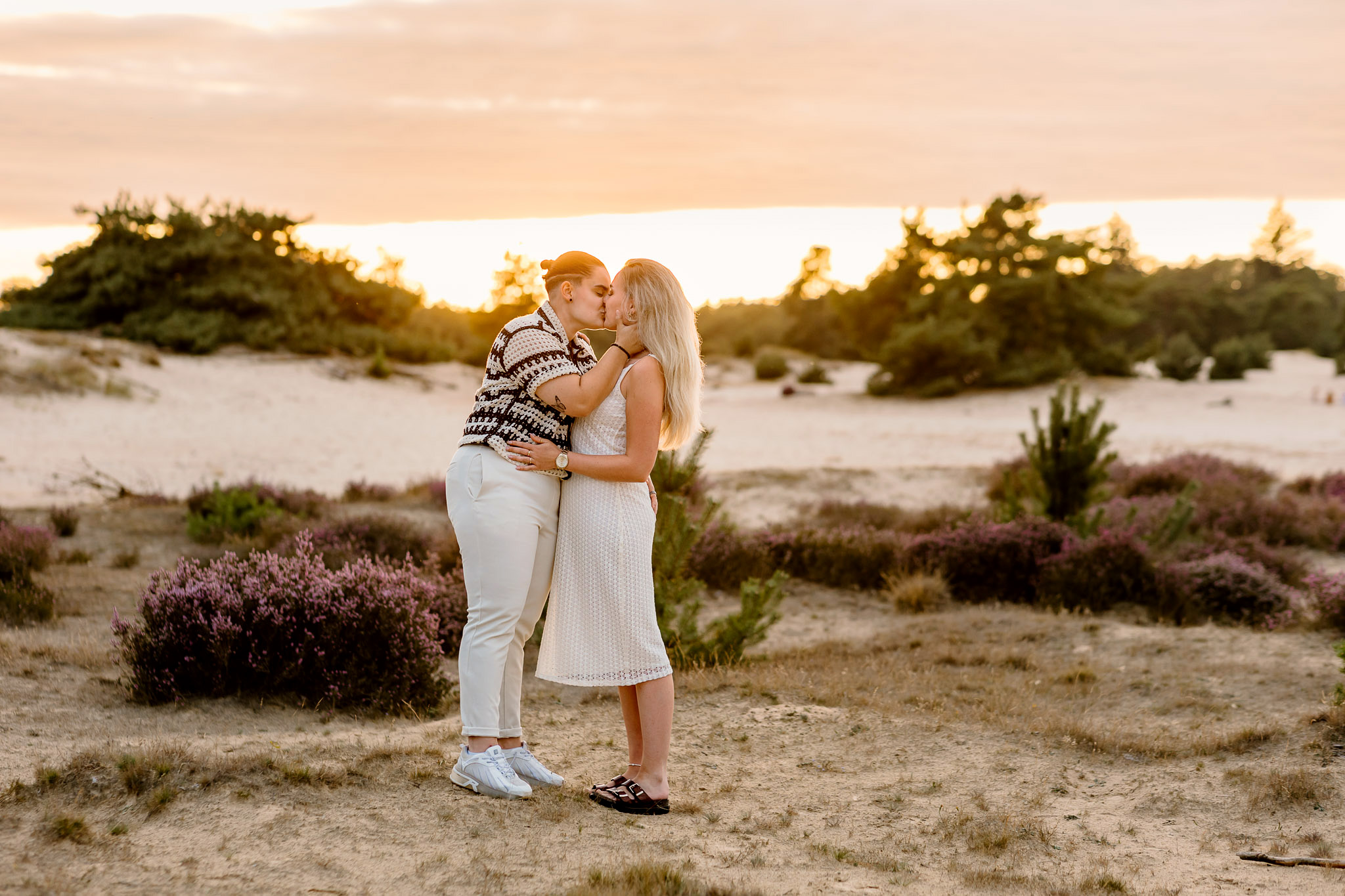 Een koppel kust in een zanderig landschap bij zonsondergang, omringd door struiken en paarse bloemen - perfecte inspiratie voor kledingtips fotoshoot ideeën.