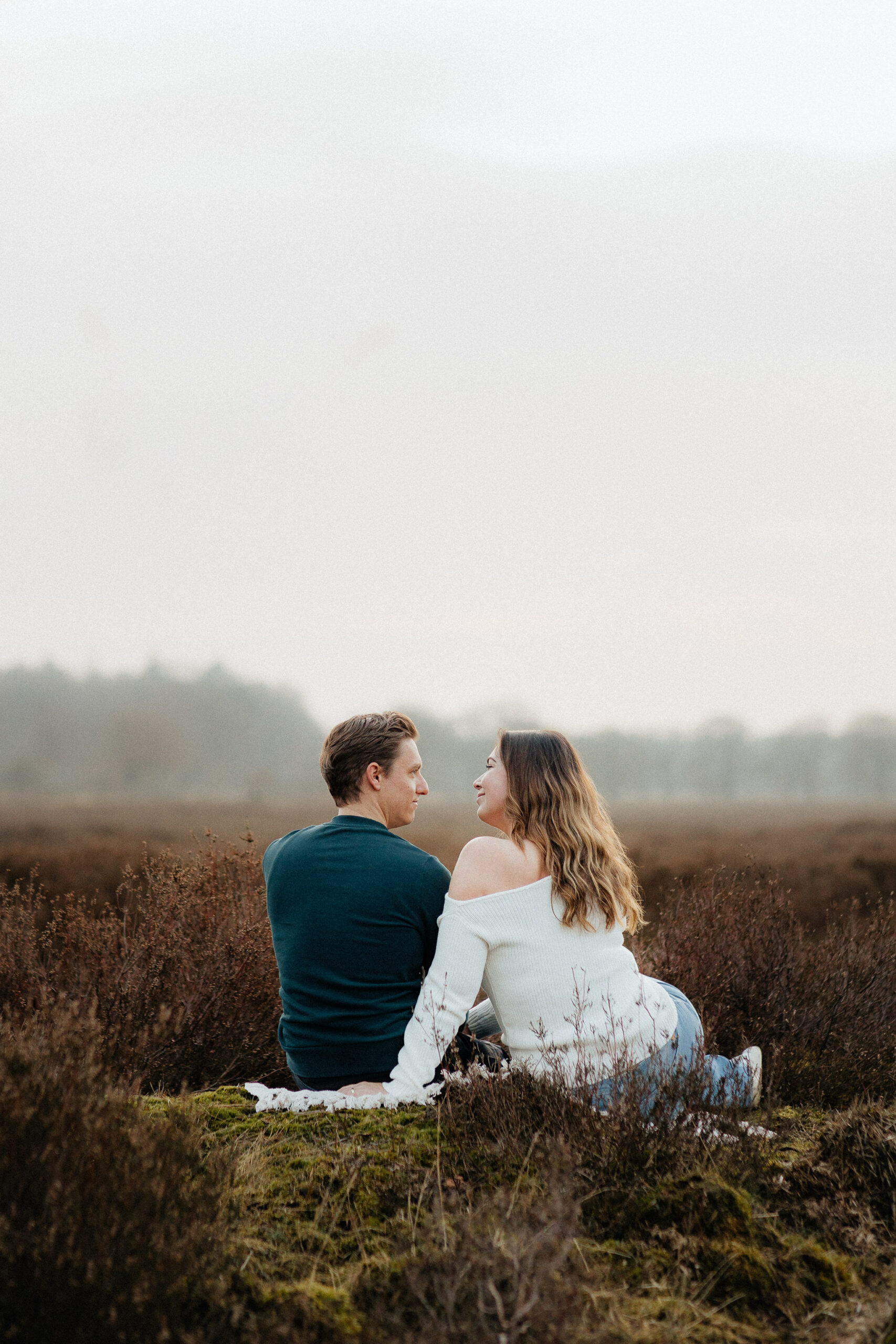 Een koppel zit buiten tussen de heide naast elkaar onder een bewolkte hemel tijdens hun koppel fotoshoot.