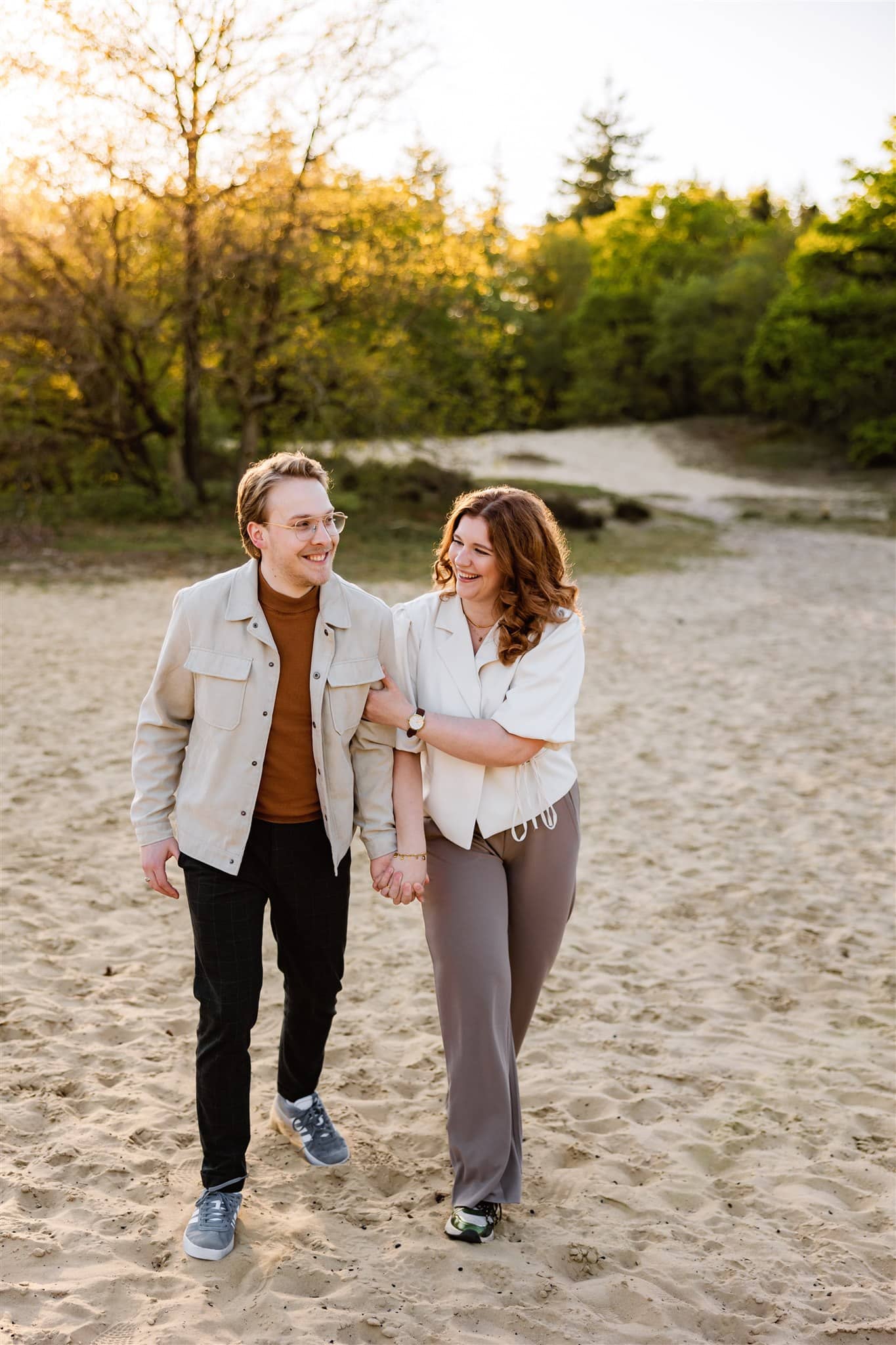 Een lachend stel loopt hand in hand over een zandpad omringd door bomen en groen, het perfecte moment vastleggend tijdens hun loveshoot.
