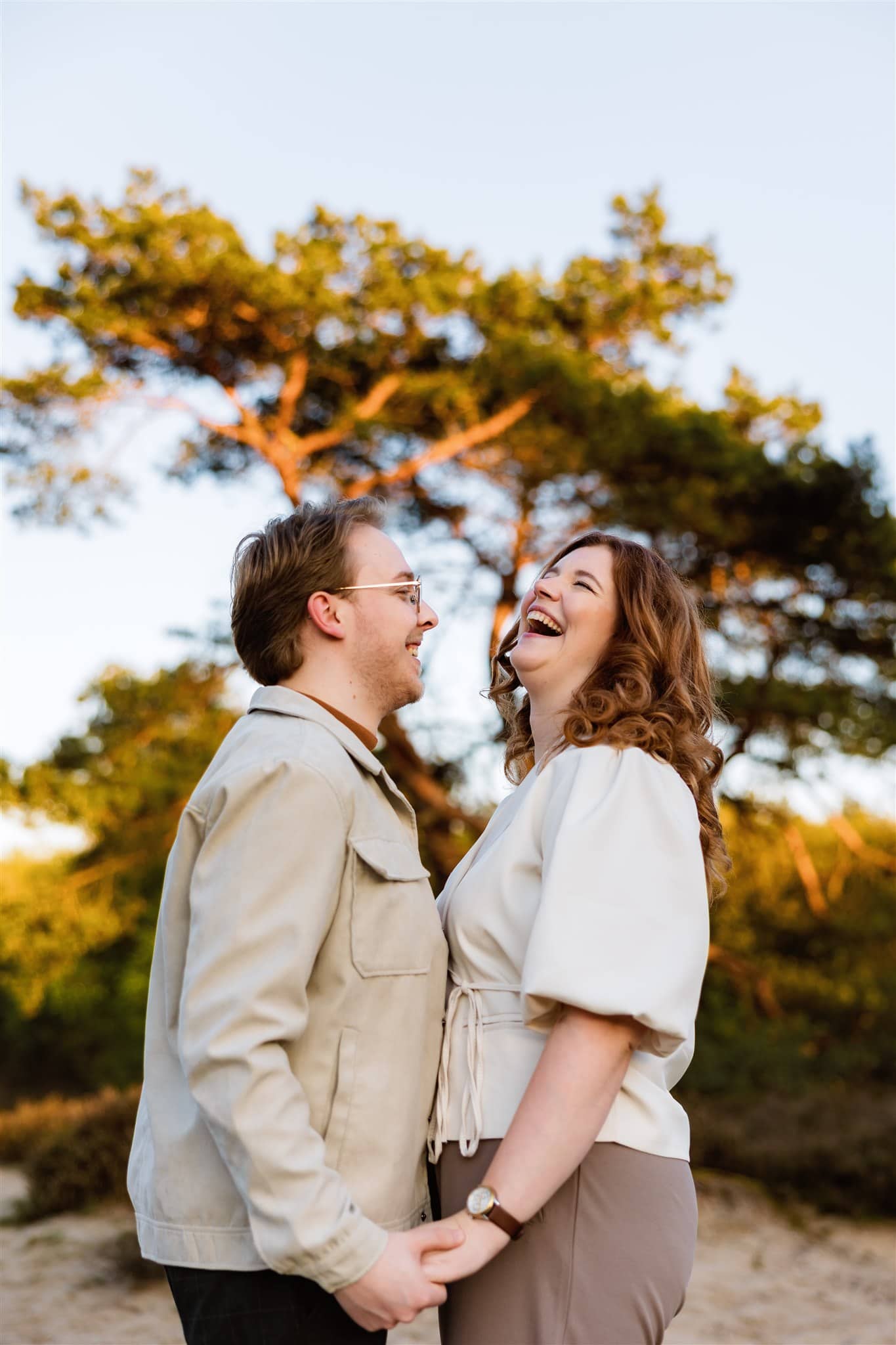 Een stel dat elkaars handen vasthoudt en samen lacht in de buitenlucht met bomen op de achtergrond tijdens een loveshoot in Groningen.
