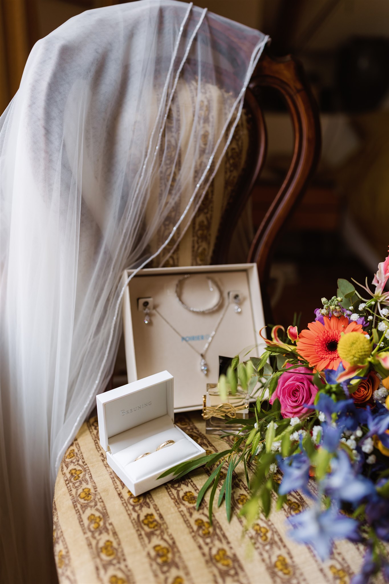 Een sluier hangt over een stoel. Op de stoel liggen de sieraden van de bruid, de trouwringen en het kleurrijke bruidsboeket.