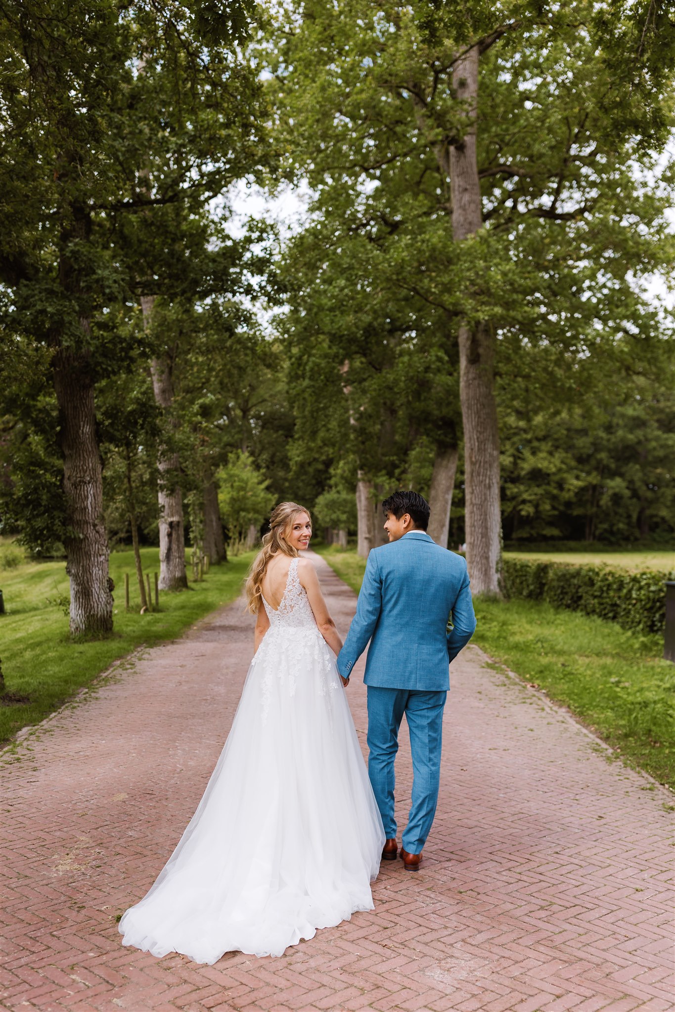 Trouwfoto van een bruidspaar dat hand in hand over de boslaan van Borg Nienoord loopt