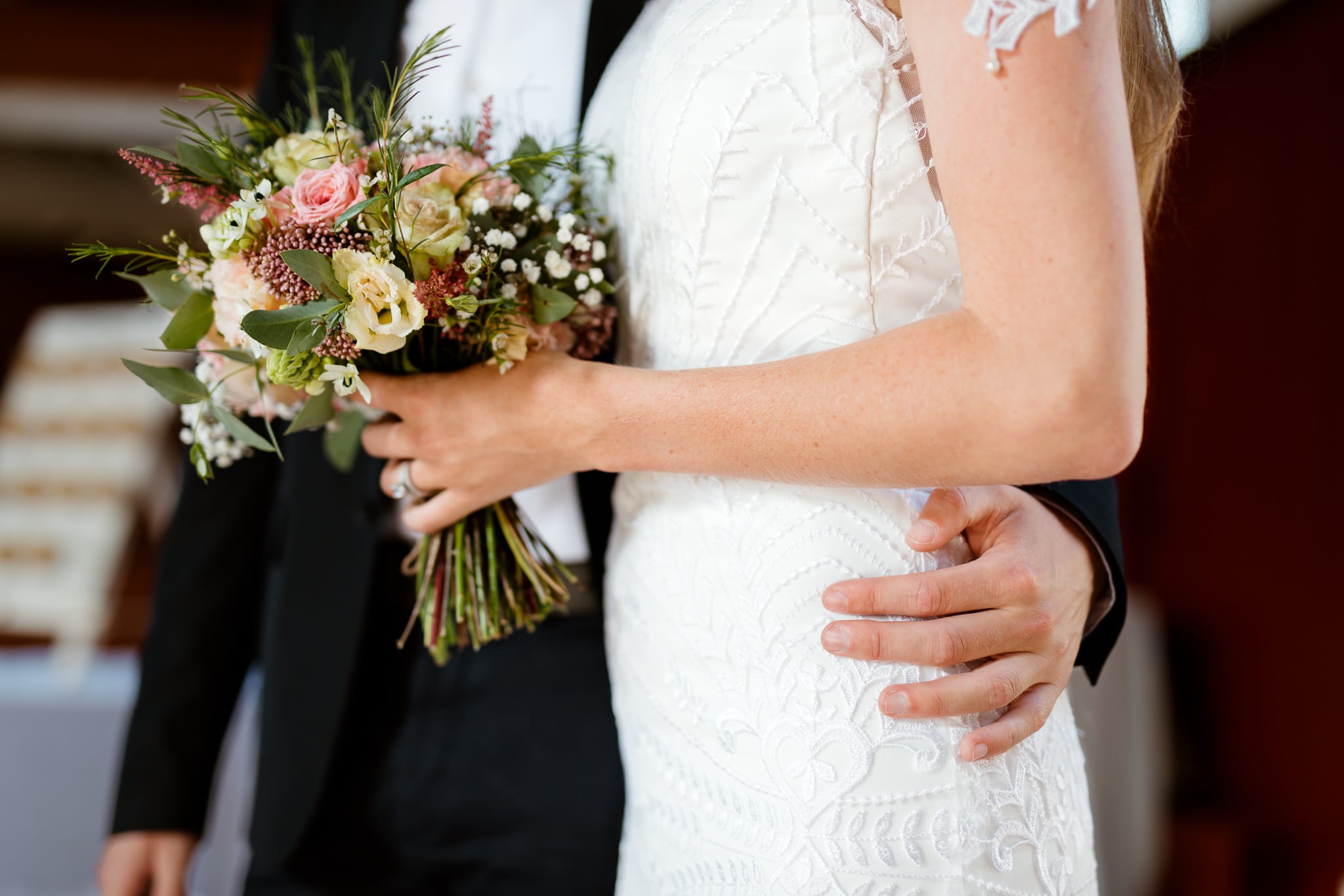 De arm van een bruidegom is om de taille van zijn bruid geslagen. De bruid in witte bruidsjurk houdt haar bloemen vast.