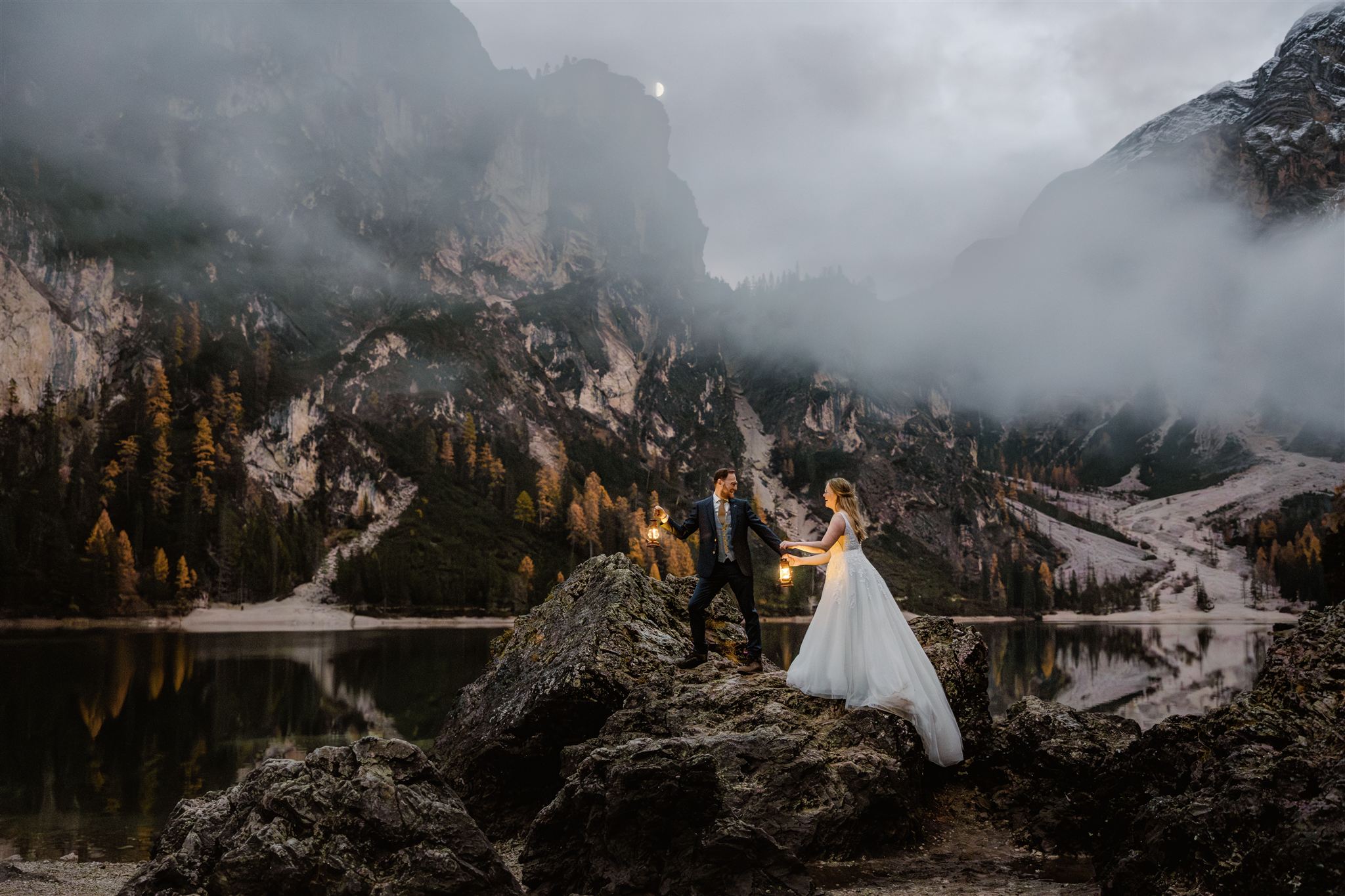 Een bruid en bruidegom staan 's avonds op een rotsachtig terrein bij bergmeer Lago di Braies in de Dolomieten en houden elkaars hand vast.