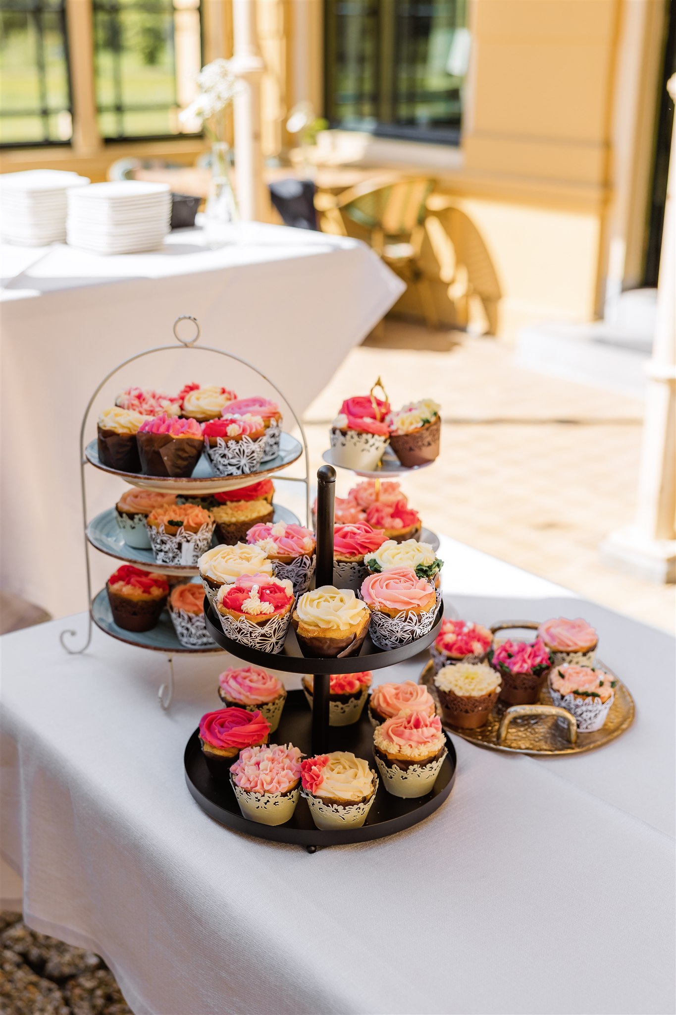 Twee cupcakestandaards met kleurrijke, met bloemen versierde cupcakes op een wit tafelkleed buiten.