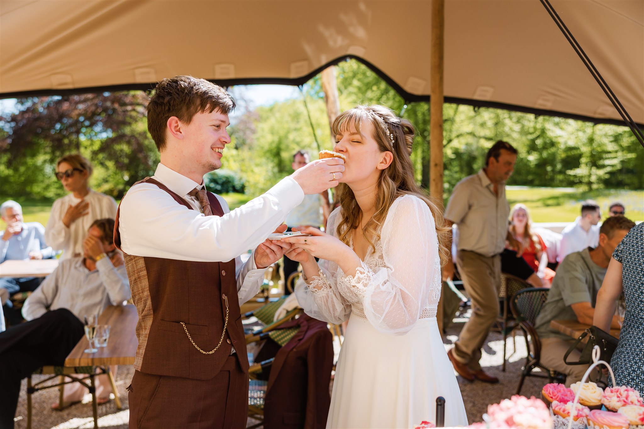 Een bruidegom voert lachend een cupcake aan een bruid op een openluchtbruiloft met gasten op de achtergrond.