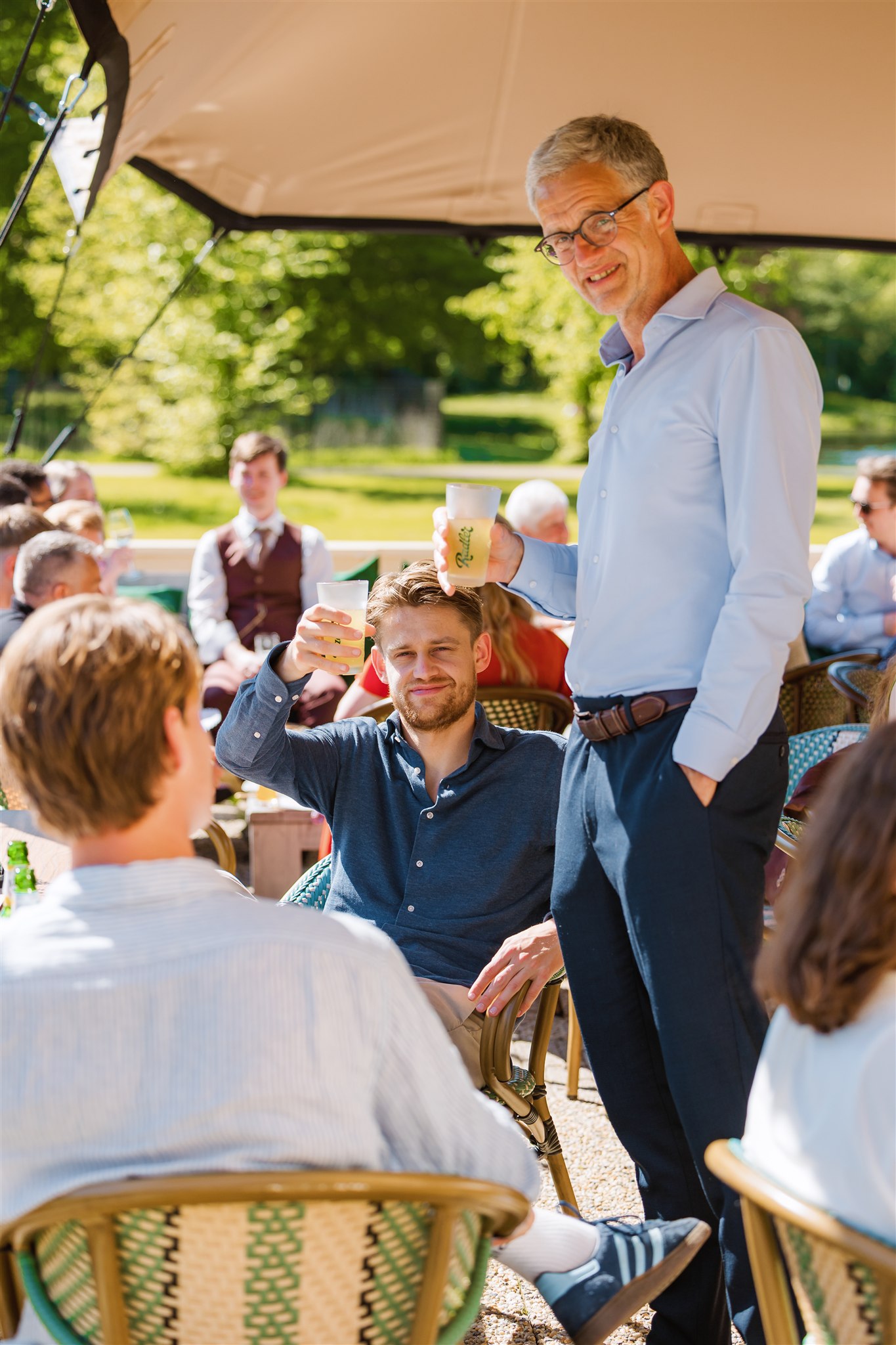 Twee mannen heffen een drankje en glimlachen tijdens de receptie van een bruiloft onder een afdak op een zonnige dag.