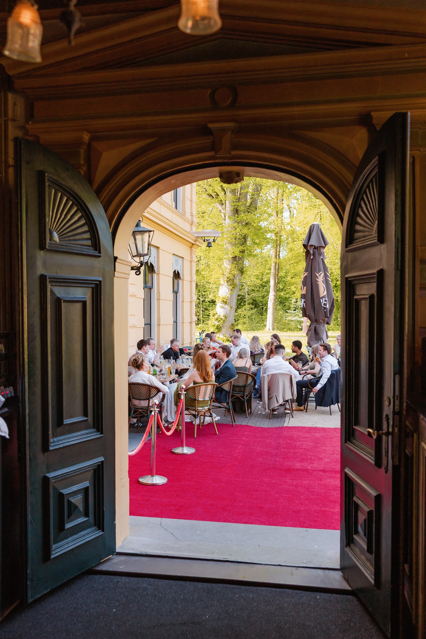 Uitzicht door open deuren op mensen die buiten borrelen op een patio met een rode loper en bomen op de achtergrond.