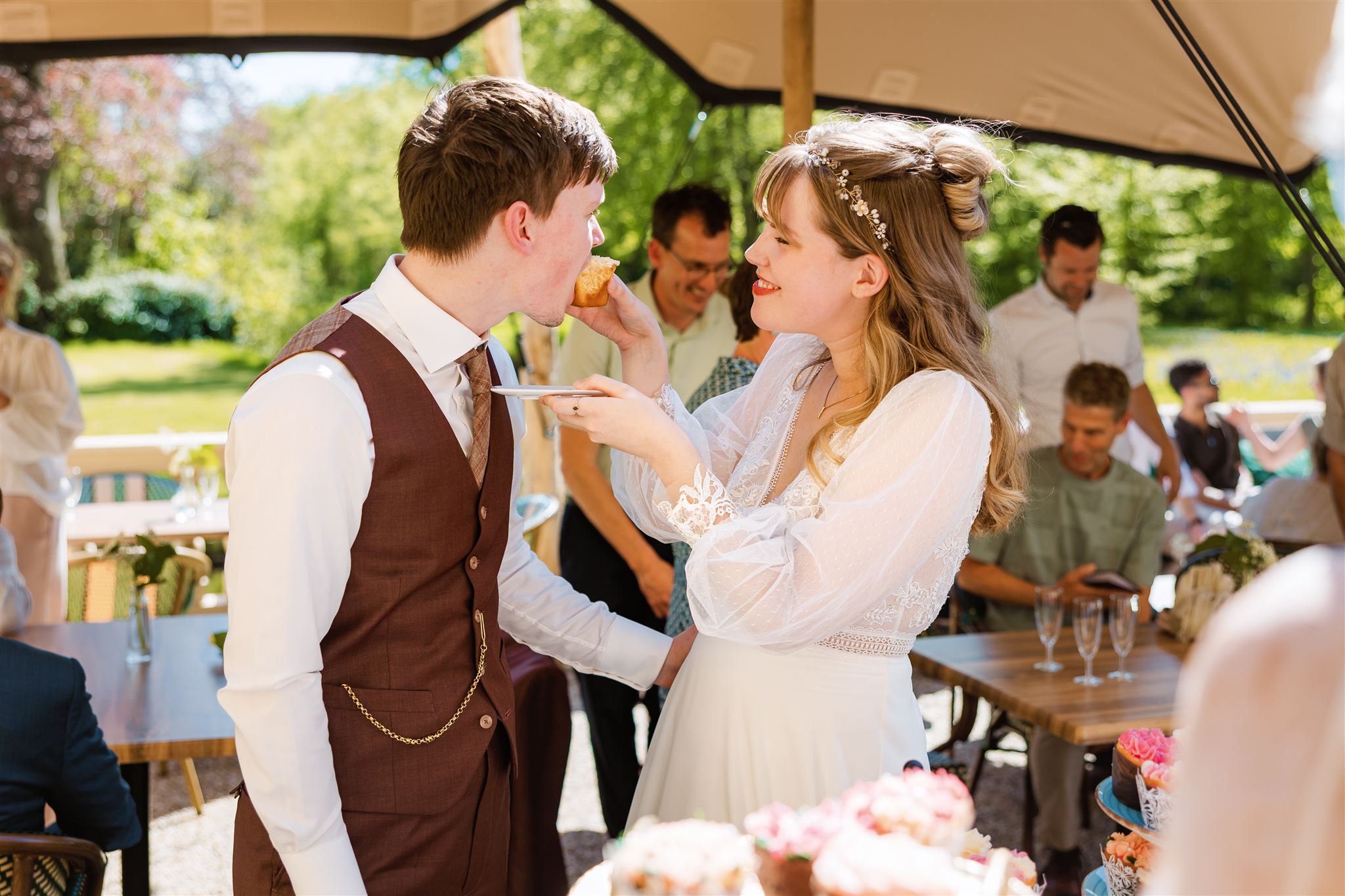 Een bruid voert de bruidegom buiten met taart tijdens een zonnig huwelijksfeest, omringd door gasten.