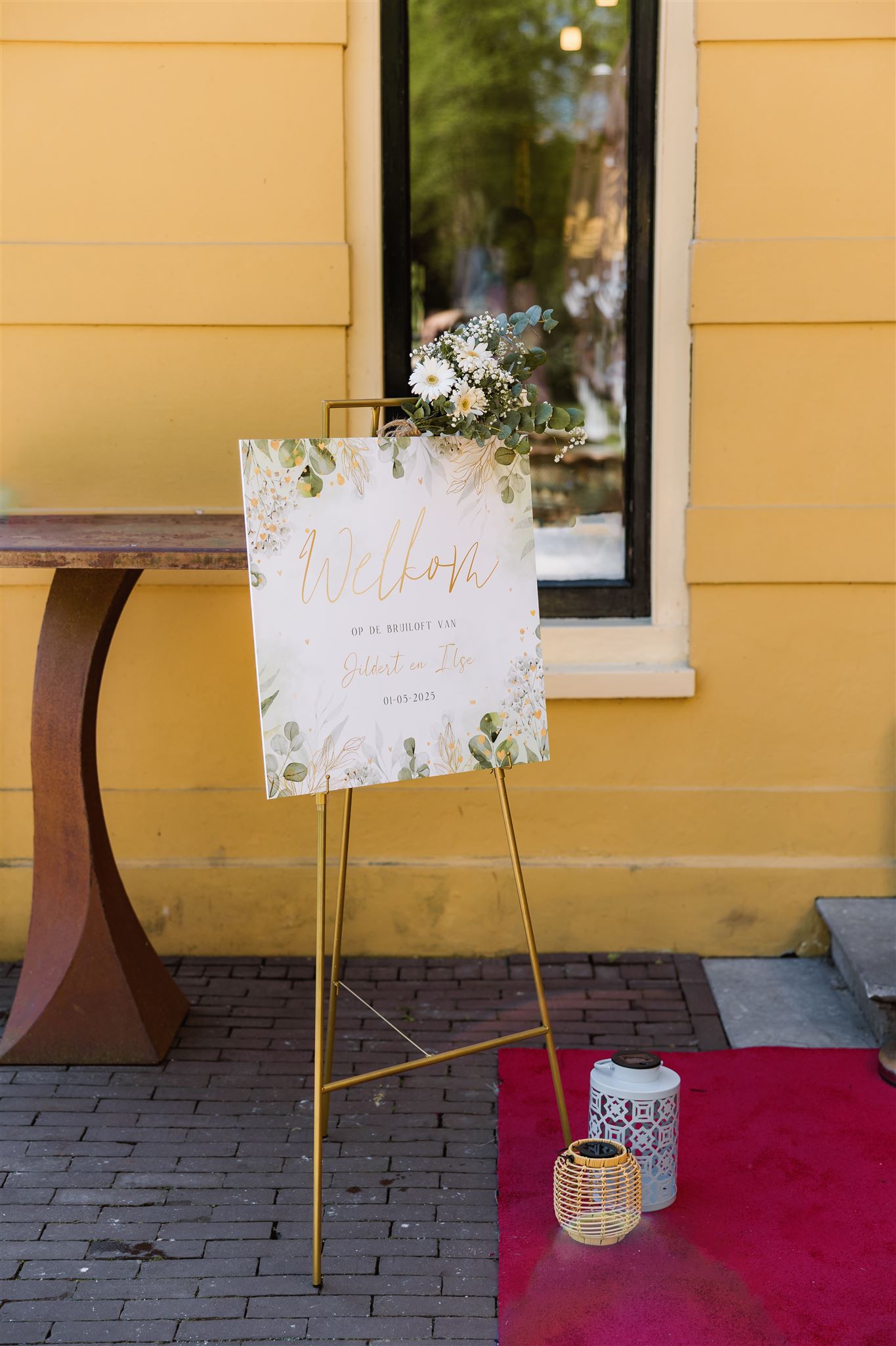 Schildersezel met een decoratief welkomstbord, bloemen en lantaarns op een rode loper buiten een geel gebouw.