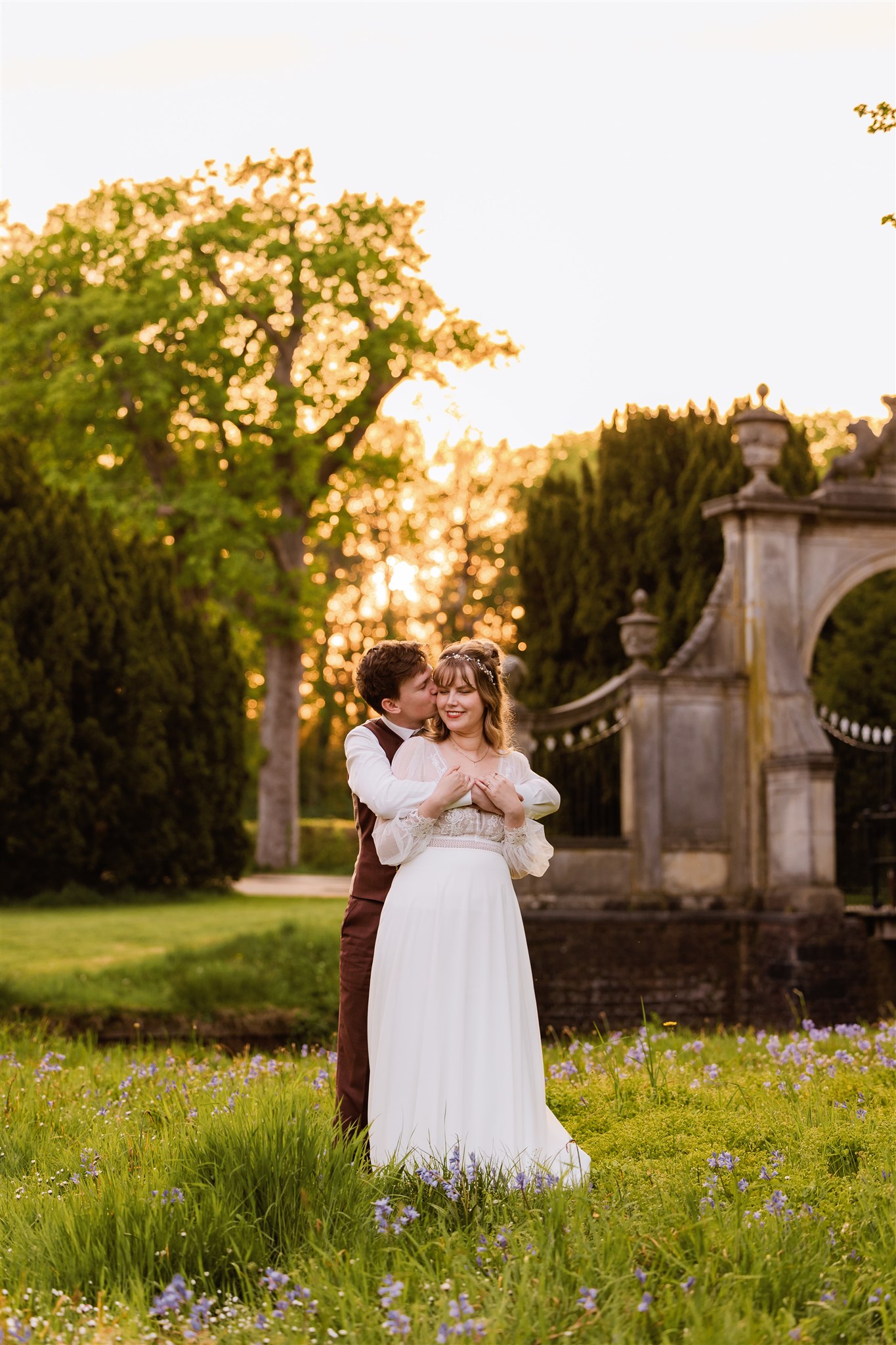 Een bruidspaar in trouwkleding omhelst elkaar in een tuin met ondergaande zon, omringd door groen en bloemen.