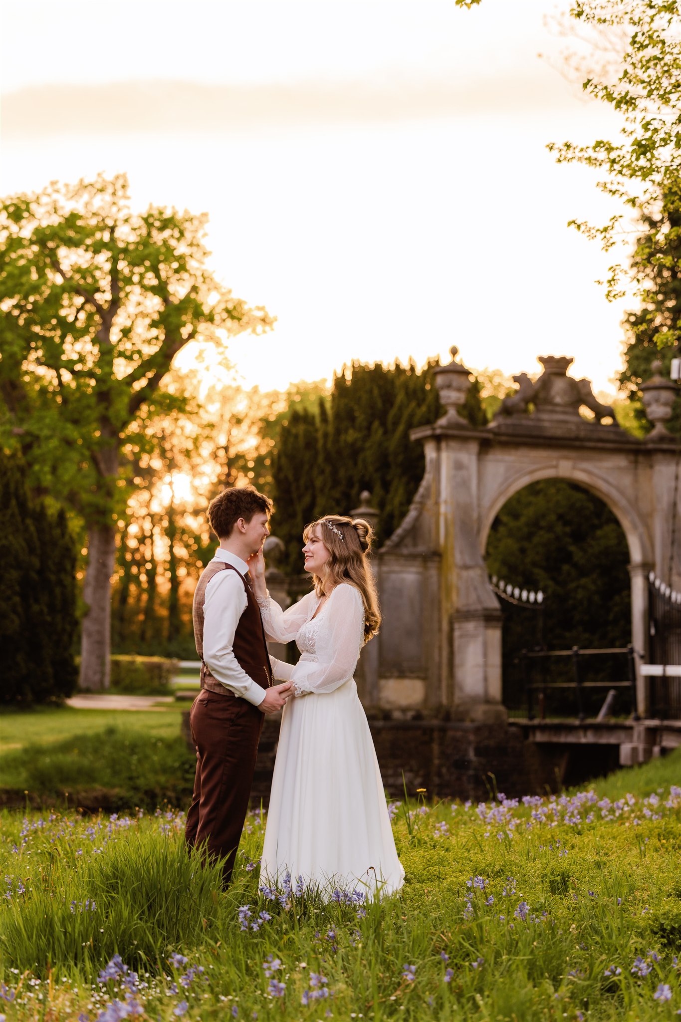 Een bruidspaar staat in de tuin voor de toegangspoort van Borg Nienoord bij zonsondergang en kijkt naar elkaar