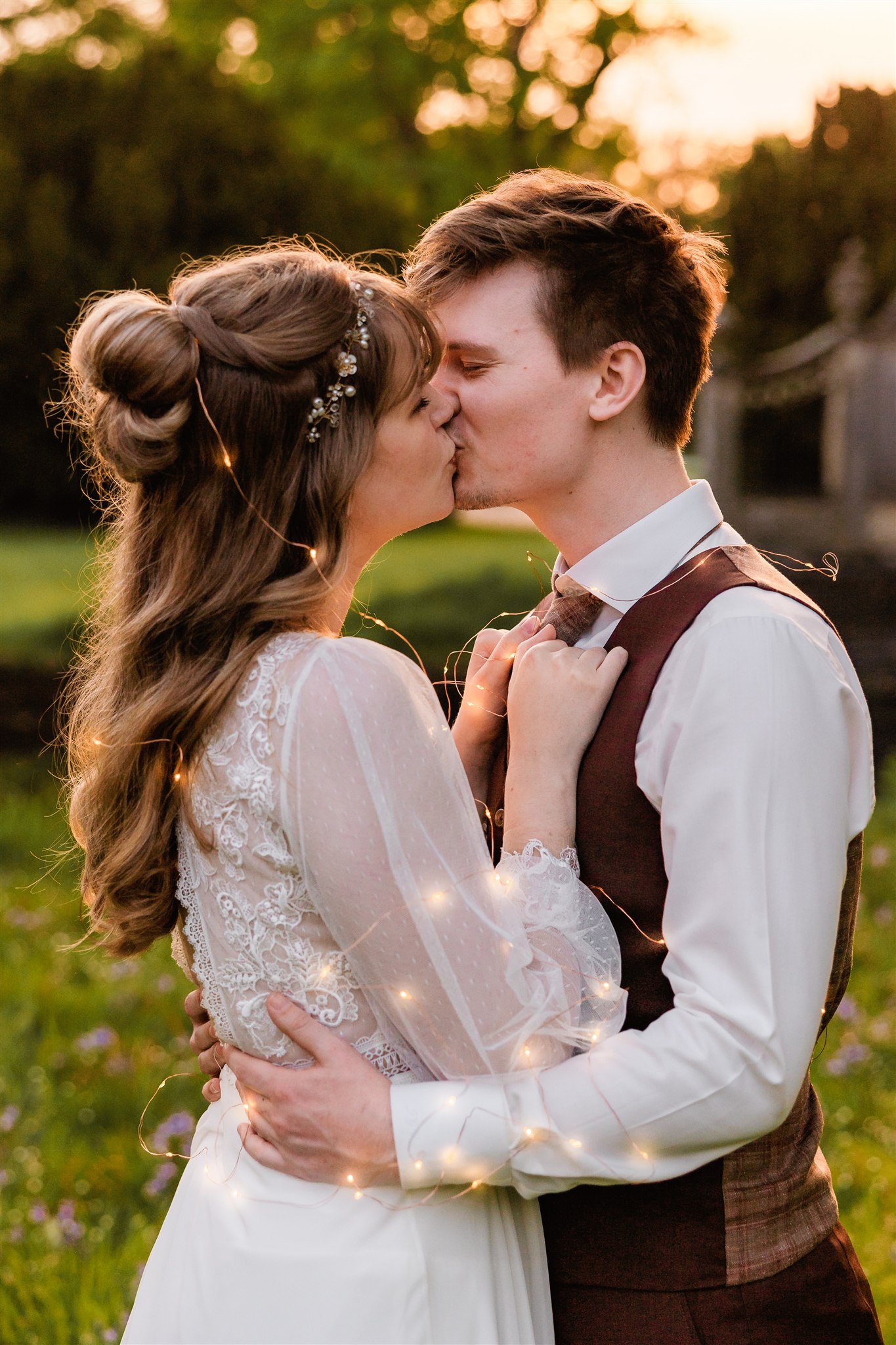 Een bruid en bruidegom kussen elkaar buiten bij zonsondergang, gehuld in gloeiende lichtjes.