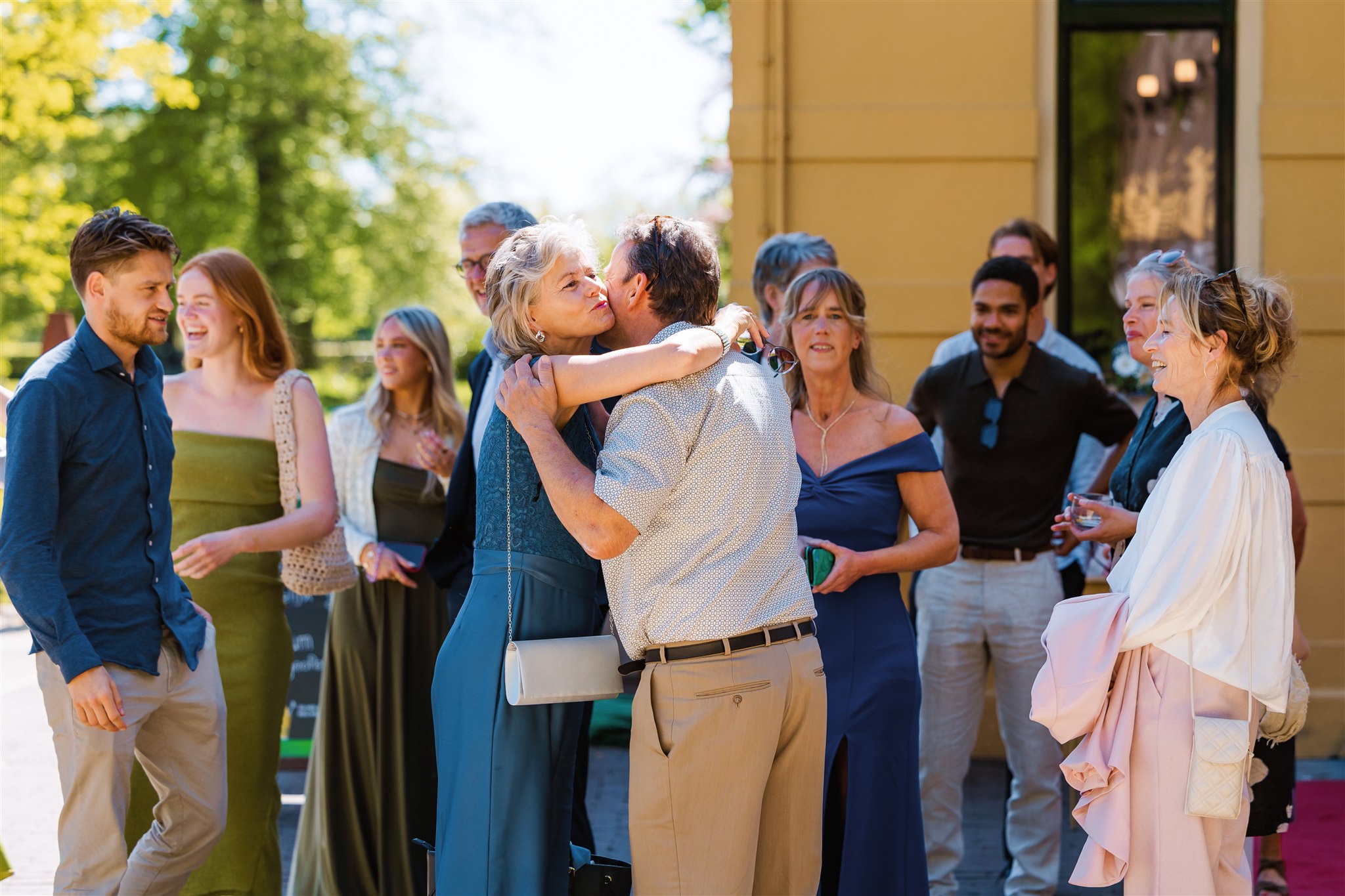 Een groep daggasten op een bruiloft in de buitenlucht, met twee mensen knuffelend en anderen lachend en pratend.