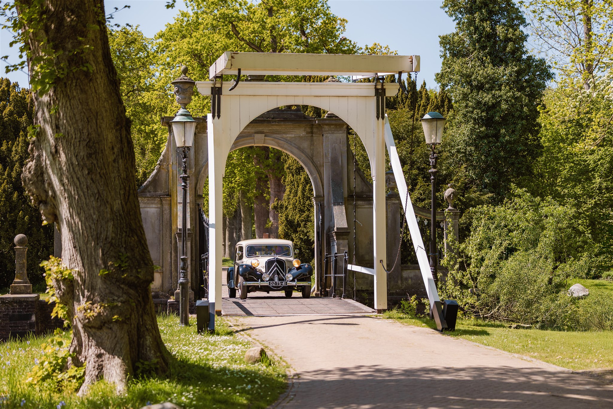 Een oldtimer rijdt over de witte ophaalbrug van Borg Nienoord omringd door groen en bomen op een zonnige dag.