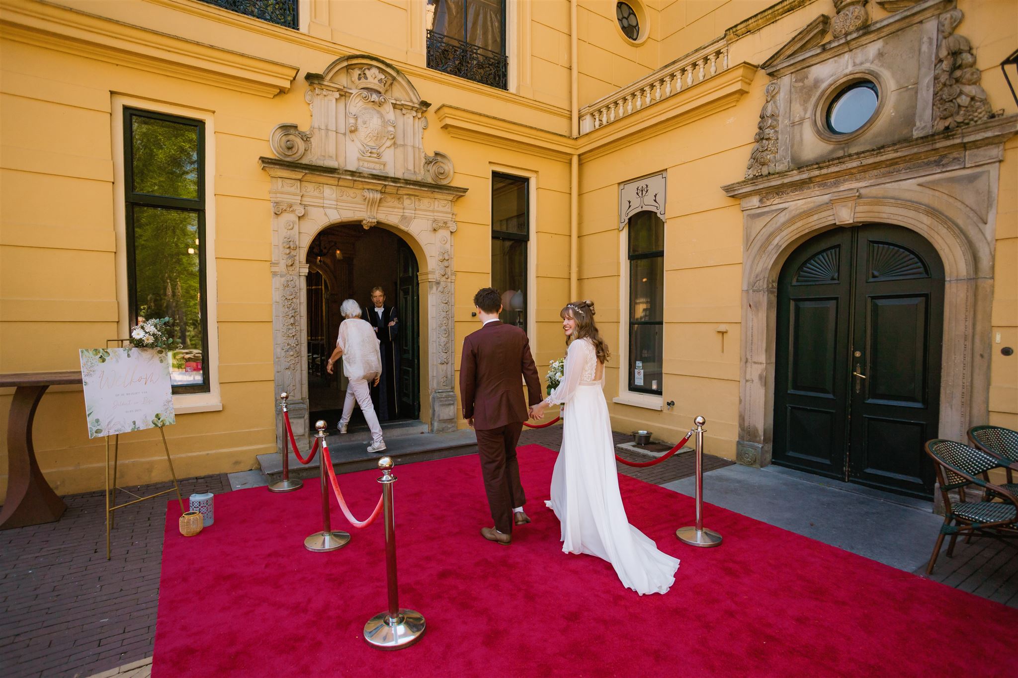 Een bruid en bruidegom lopen over een rode loper naar de ingang van een historisch geel gebouw.