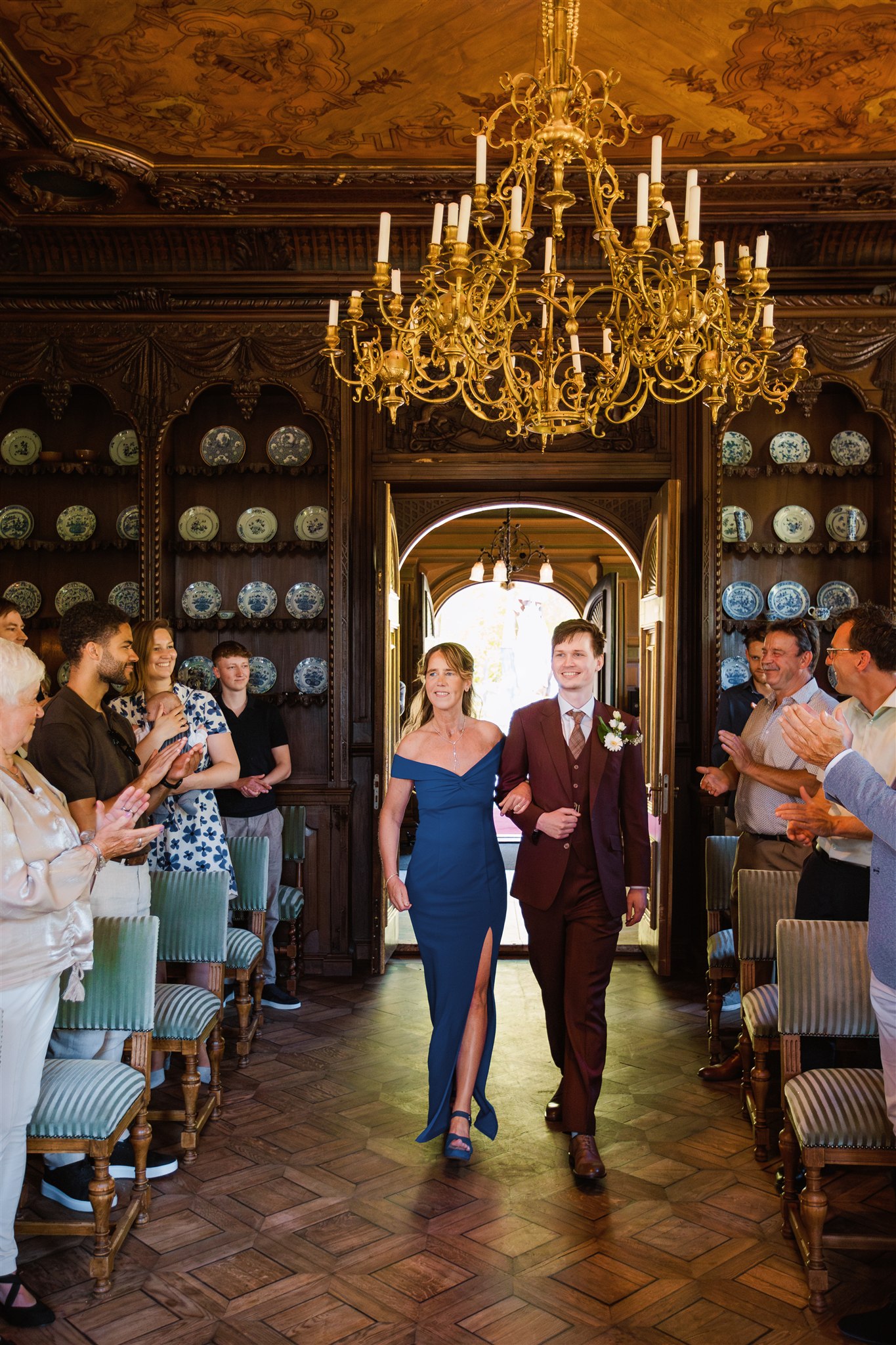 Bruidegom loopt met zijn moeder door het gangpad onder een gouden kroonluchter in de Ridderzaal terwijl gasten applaudisseren.