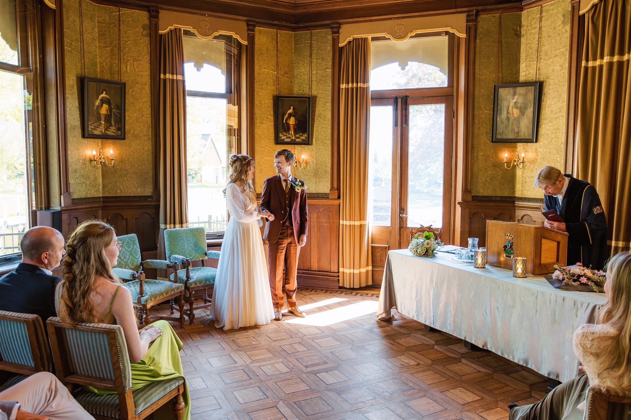 Een bruid en bruidegom houden elkaars hand vast tijdens een huwelijksceremonie in een zonovergoten, elegante kamer met zittende gasten.
