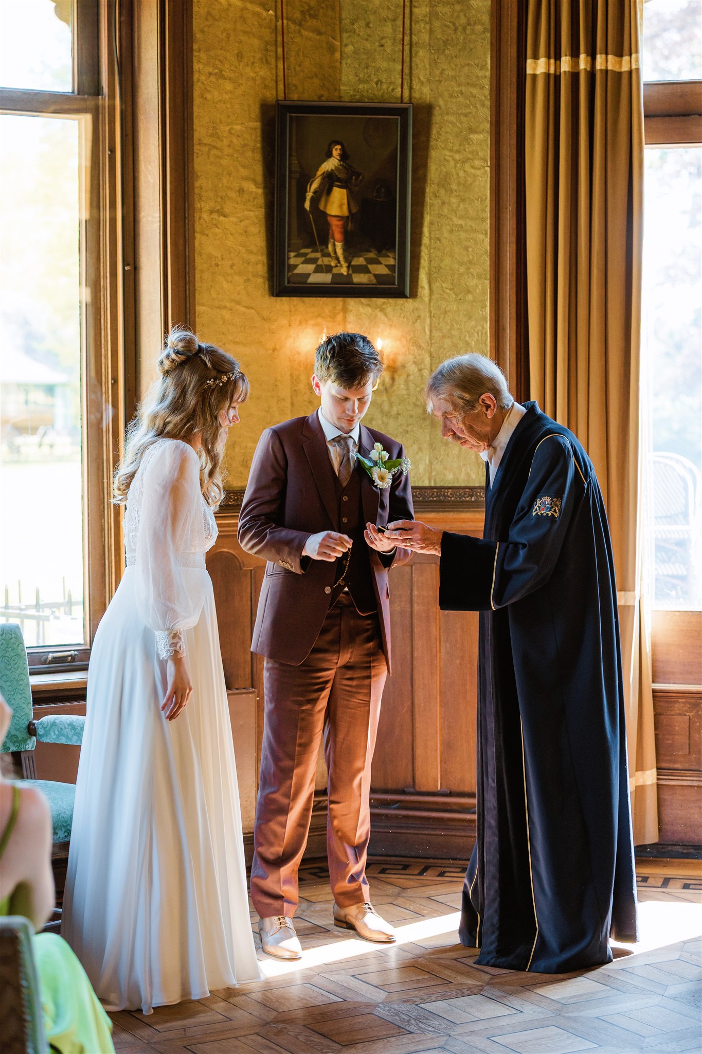 Een koppel wisselt ringen uit tijdens een huwelijksceremonie met de trouwambtenaar in een zonovergoten kamer met houten panelen.
