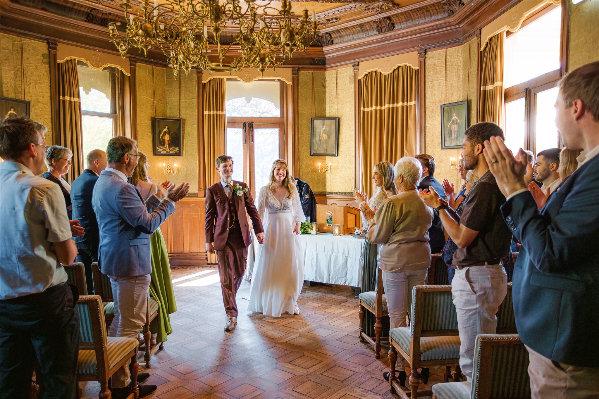 Een pasgetrouwd stel loopt door het gangpad terwijl gasten staan te applaudisseren in de elegante Ridderzaal.
