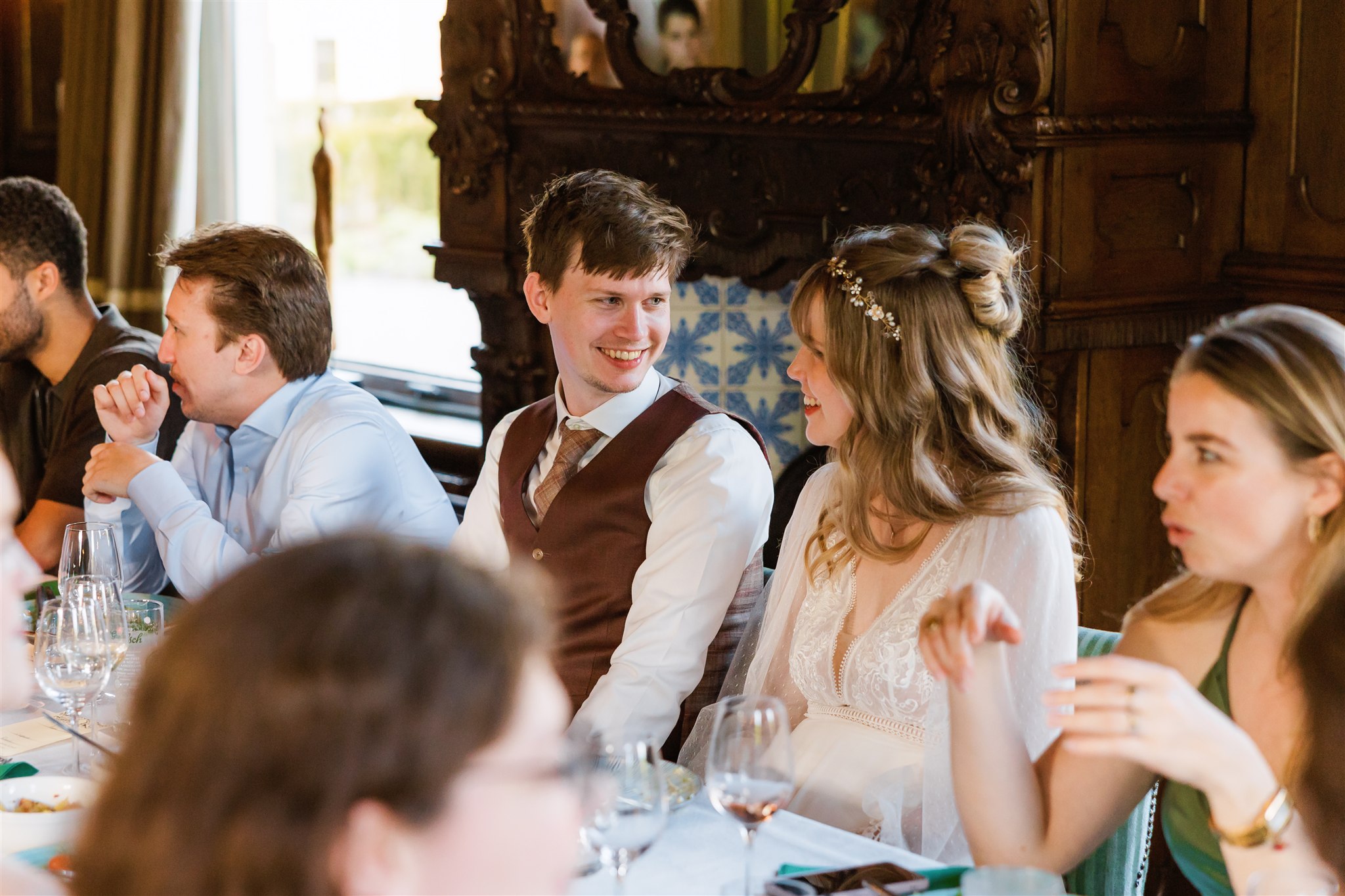 Een bruid en bruidegom lachen naar elkaar terwijl ze aan een tafel zitten met gasten tijdens het diner