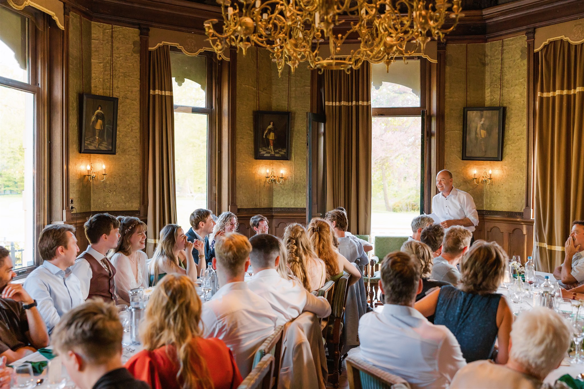 Een groep bruiloftsgasten aan tafels in de Ridderzaal van Borg Nienoord, luisterend naar de vader van de bruid die een toespraak houdt.