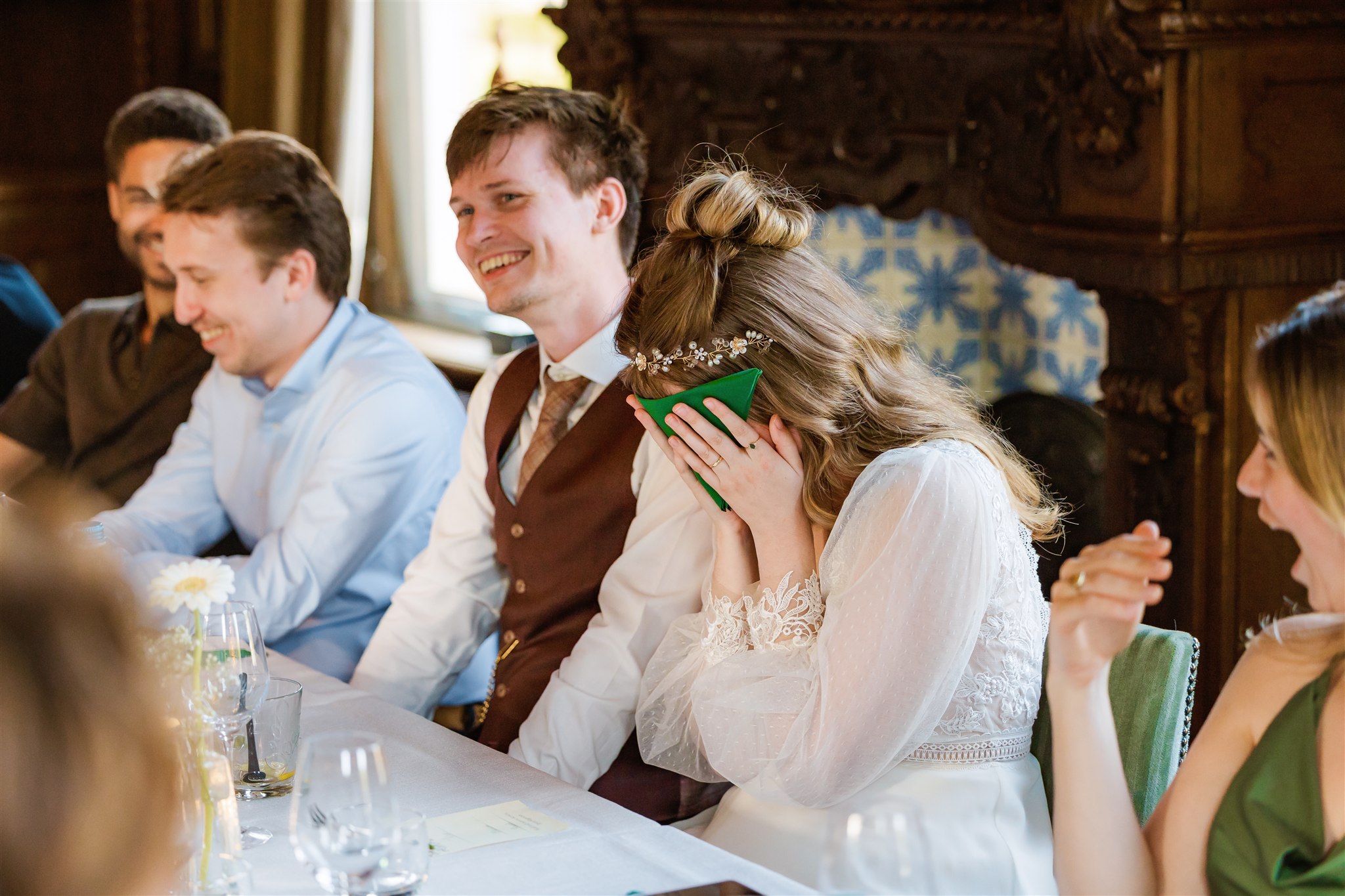 Een bruid lacht en bedekt haar gezicht met een servet uit schaamte, omringd door lachende gasten aan een gedekte eettafel.