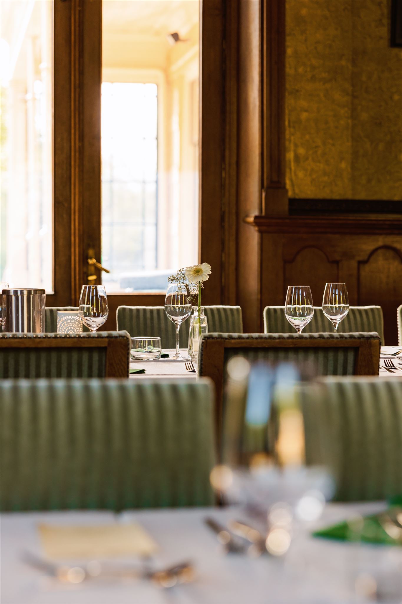 Elegante eettafel gedekt met glaswerk en bloemen in een zonovergoten, klassieke kamer met groene stoelen.