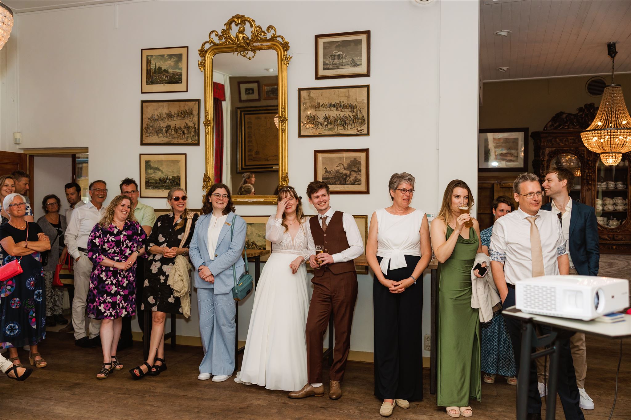 Een groep mensen, waaronder een bruidspaar, staat lachend naar iets te kijken op een binnenfeest.