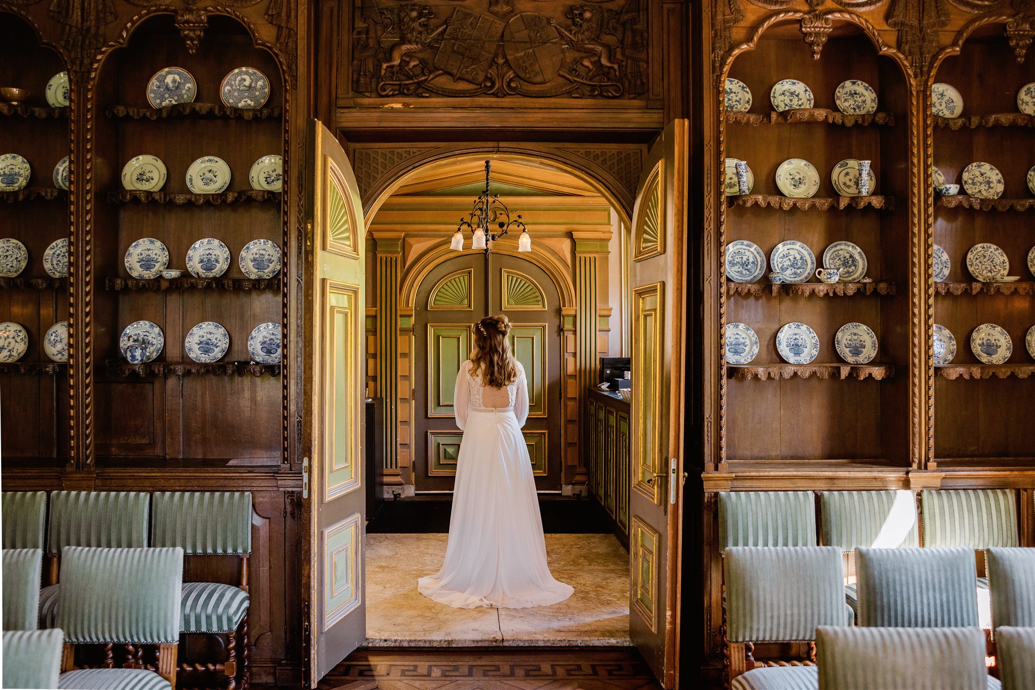 Een bruid in een witte jurk staat alleen in een grote, sierlijke kamer met planken vol decoratieve borden.
