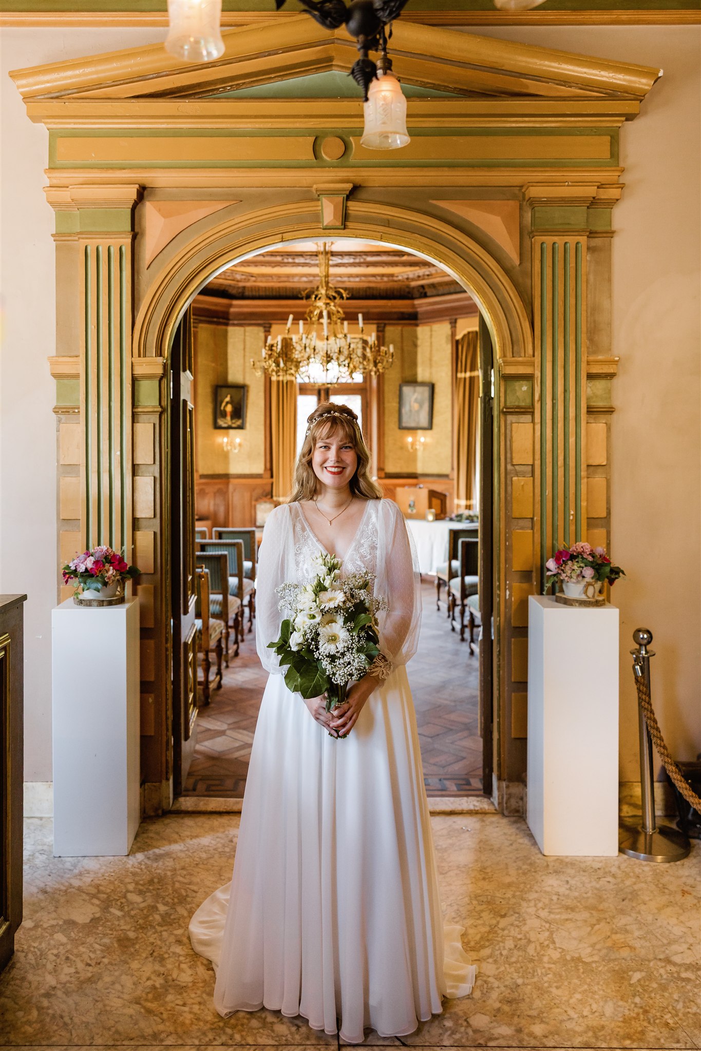 Glimlachende bruid in een witte jurk met een boeket in een sierlijke, elegante kamer met gouden accenten.