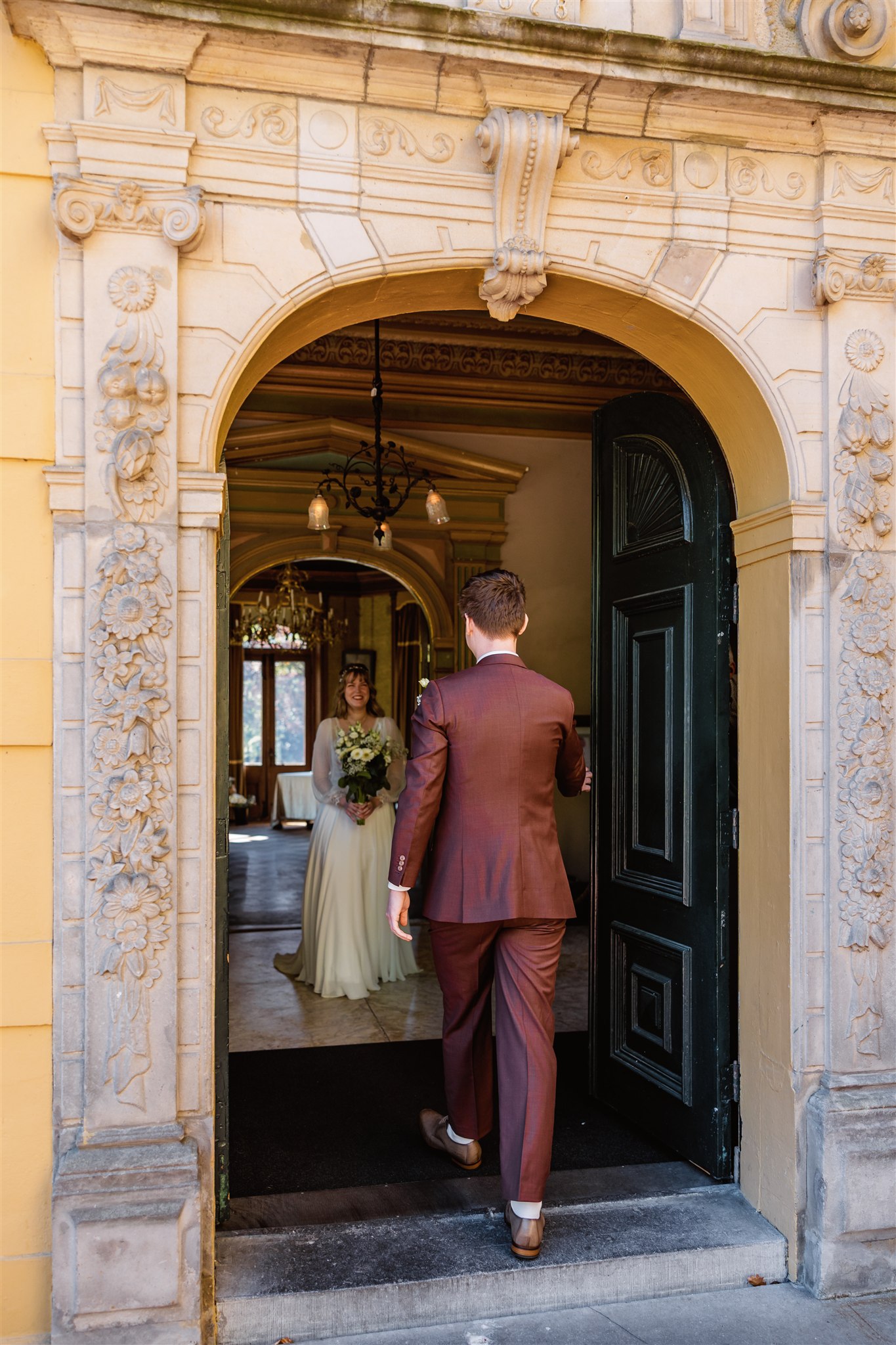 Een bruidegom gaat een grote deuropening binnen en ziet de bruid binnen staan met een boeket in haar hand tijdens hun first look.