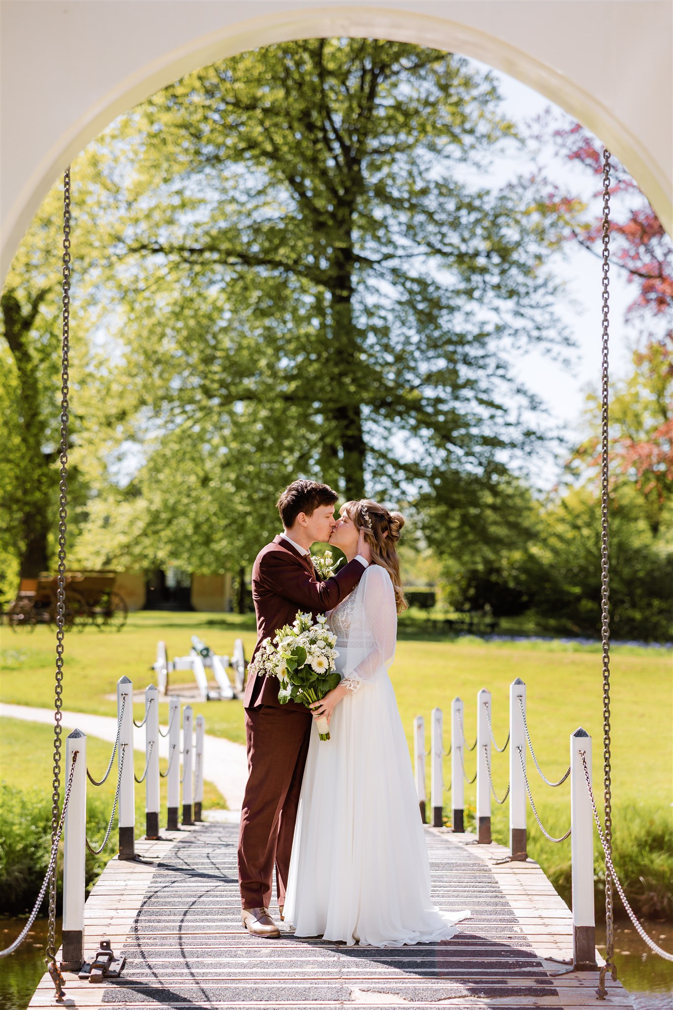 Een bruid en bruidegom kussen elkaar op een bruggetje onder een boog, omringd door groen en zonlicht.