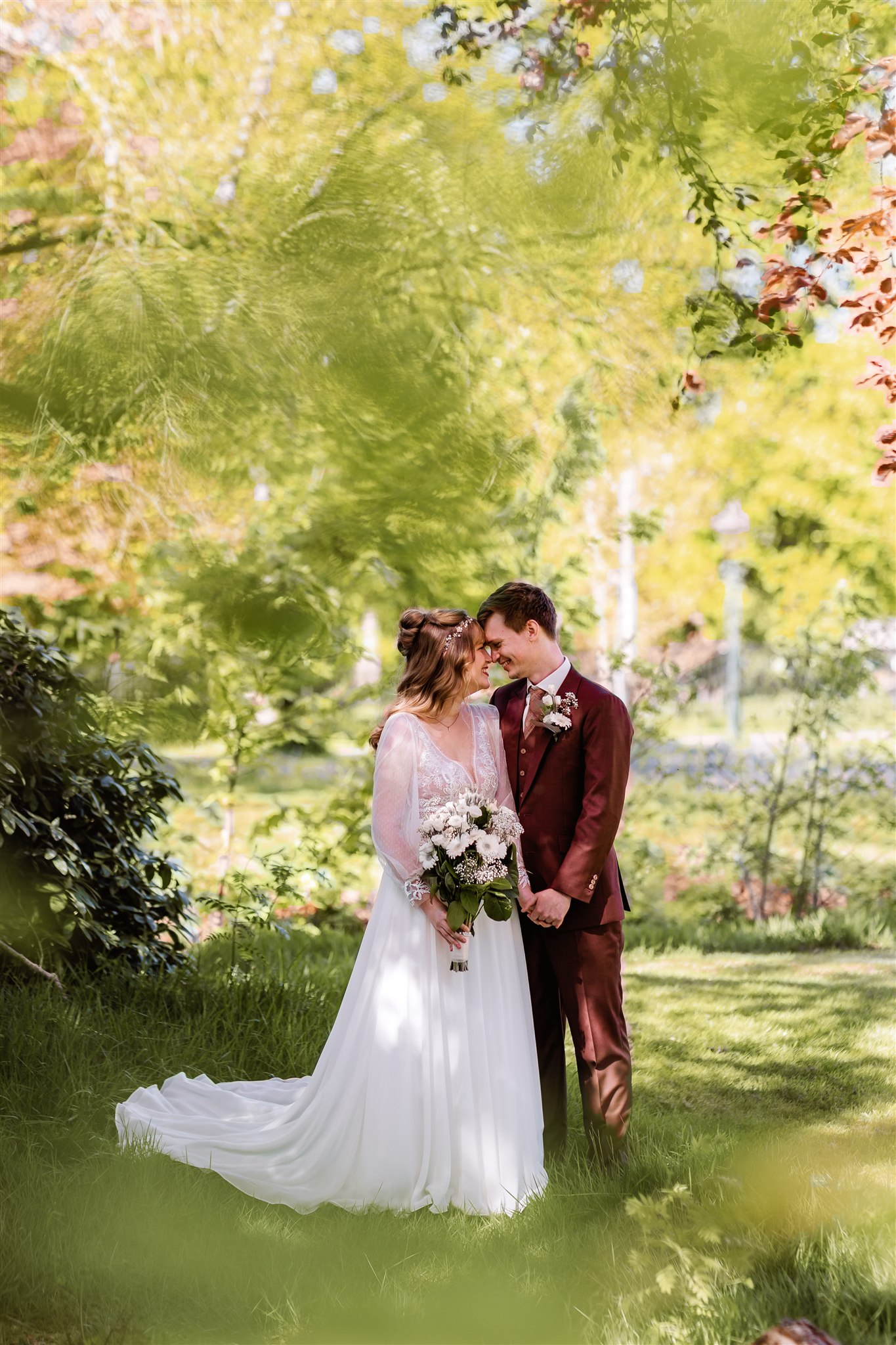 Bruid met een boeket in de hand en bruidegom staan dicht bij elkaar in een zonovergoten tuin, omringd door groen.