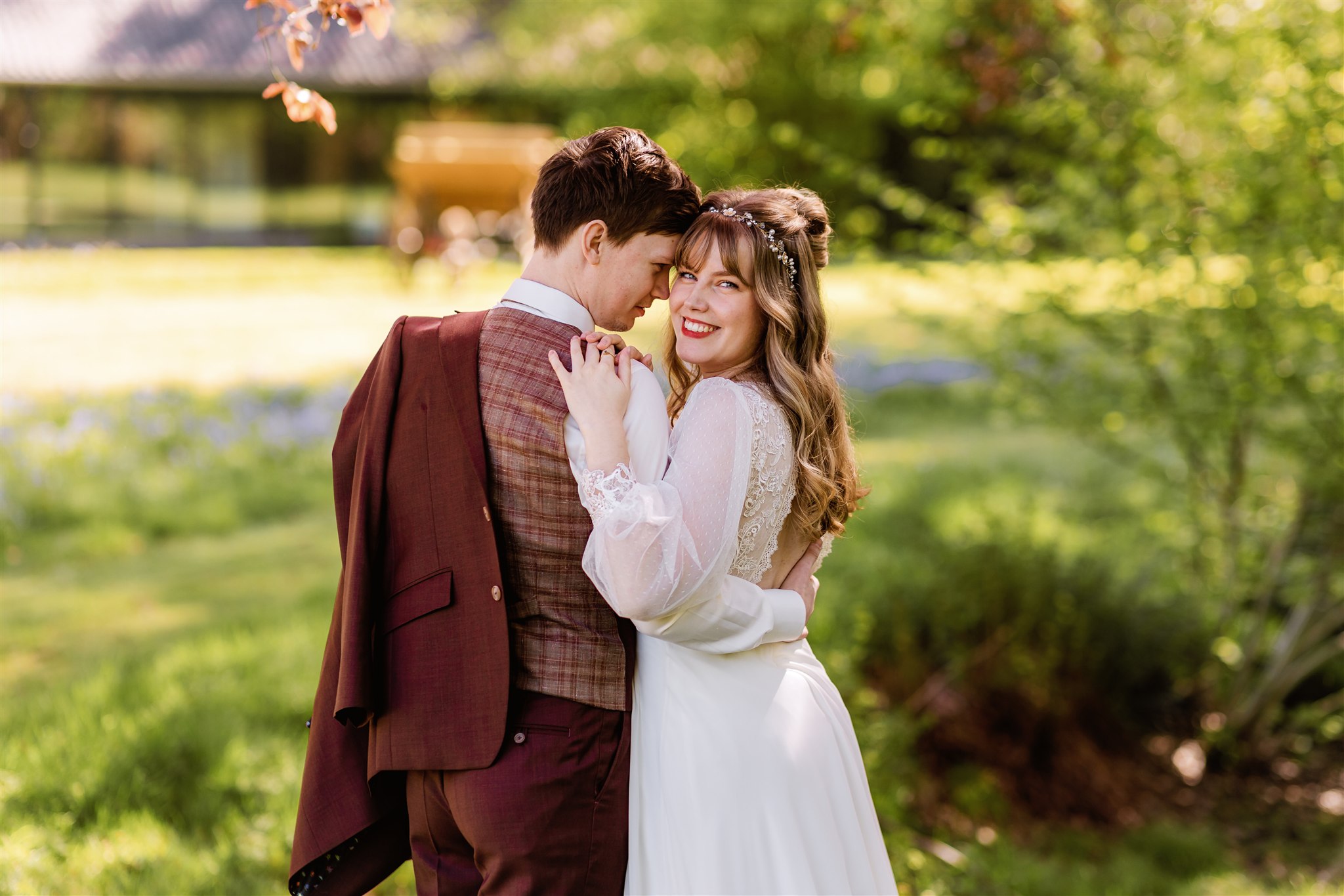 Een lachende bruid in een witte jurk kijkt in de camera en omhelst haar partner buiten op een zonnige, groene dag.