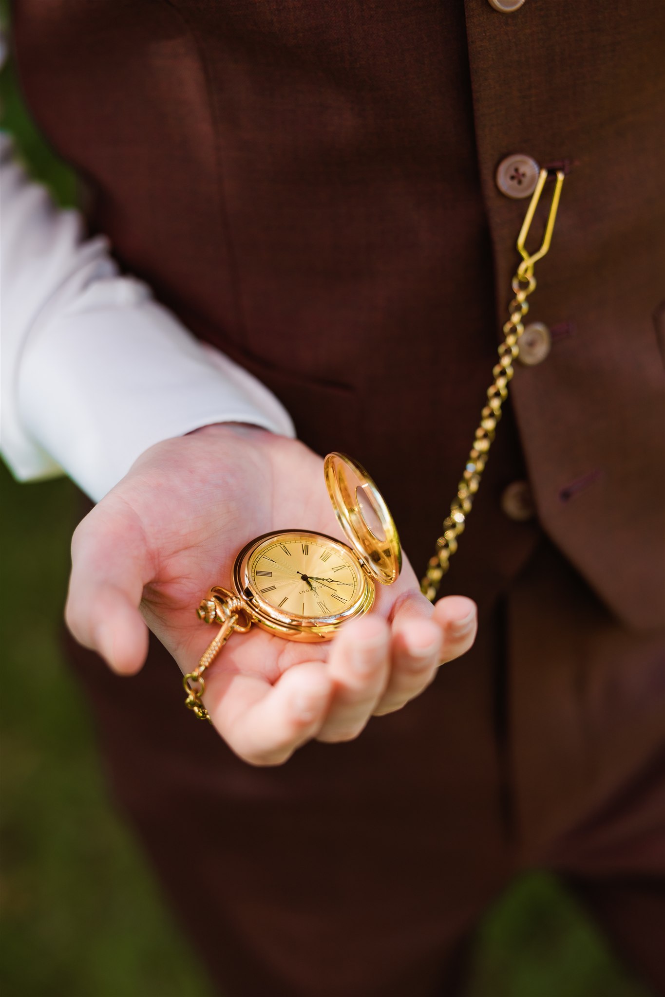 Bruidegom in een bordeaux pak houdt een open gouden zakhorloge aan een ketting in zijn hand.