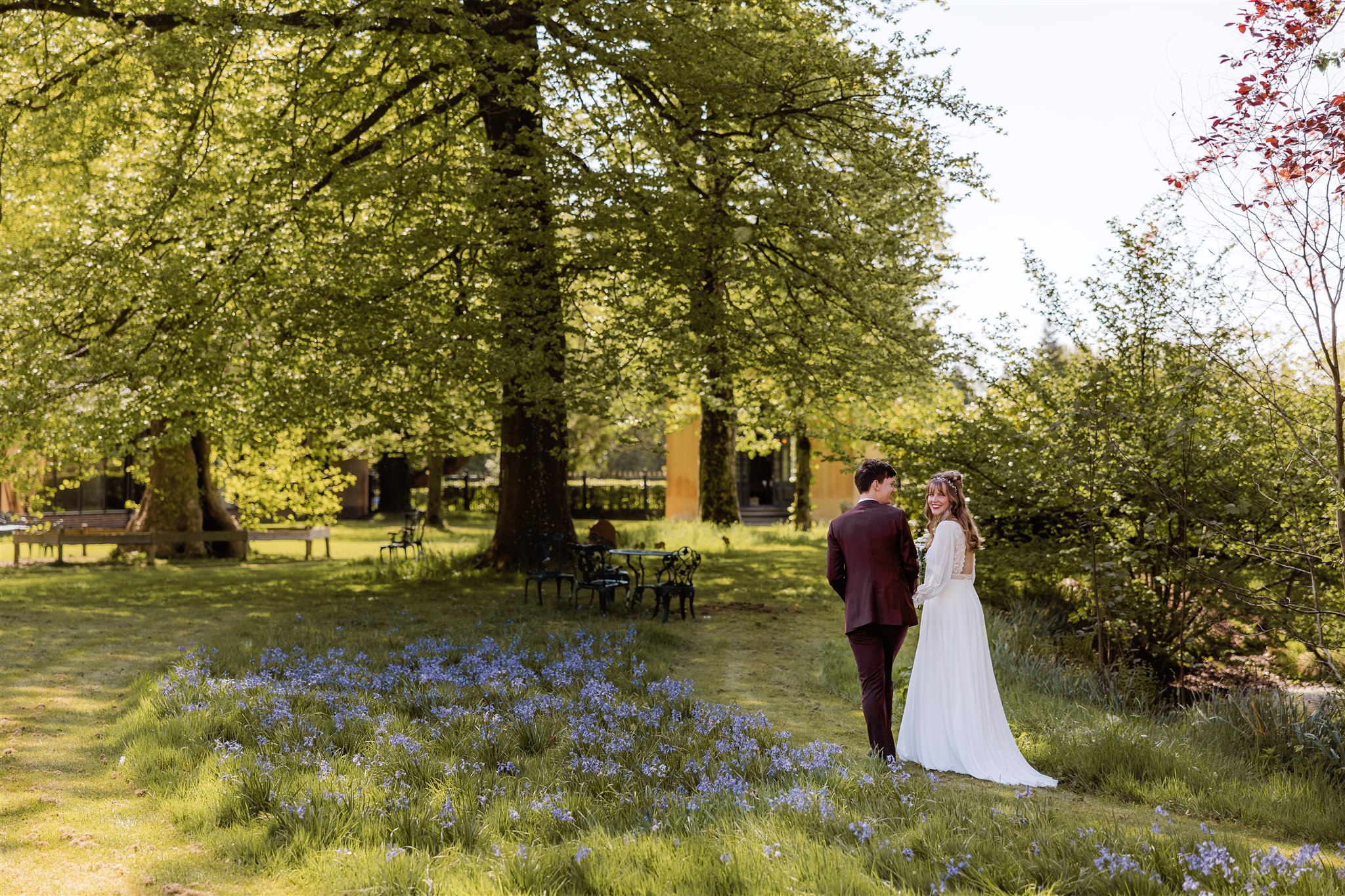 Een bruid en bruidegom staan samen in de zonovergoten tuin van Borg Nienoord met bloeiende blauwe bloemen en hoge groene bomen.